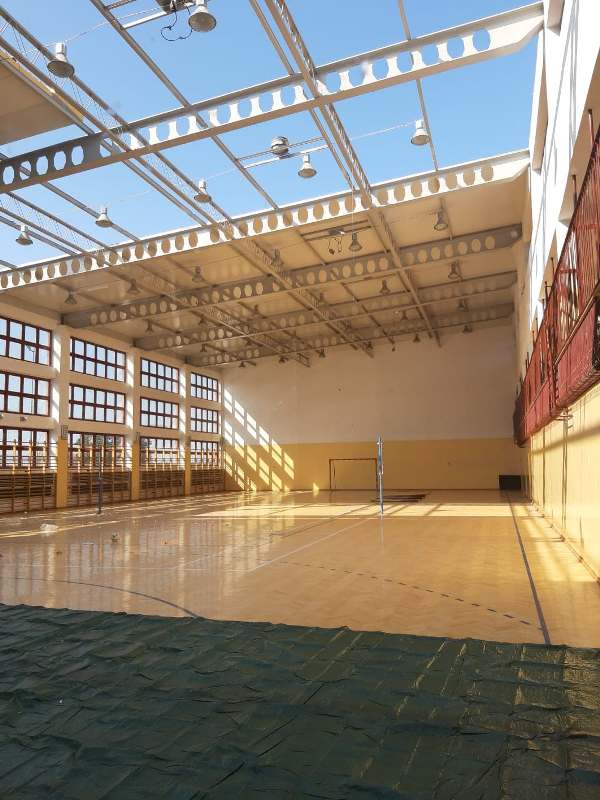 Wnętrze hali sportowej z dużymi oknami, drewnianą podłogą, siatką do siatkówki i metalową konstrukcją dachu z żarówkami.