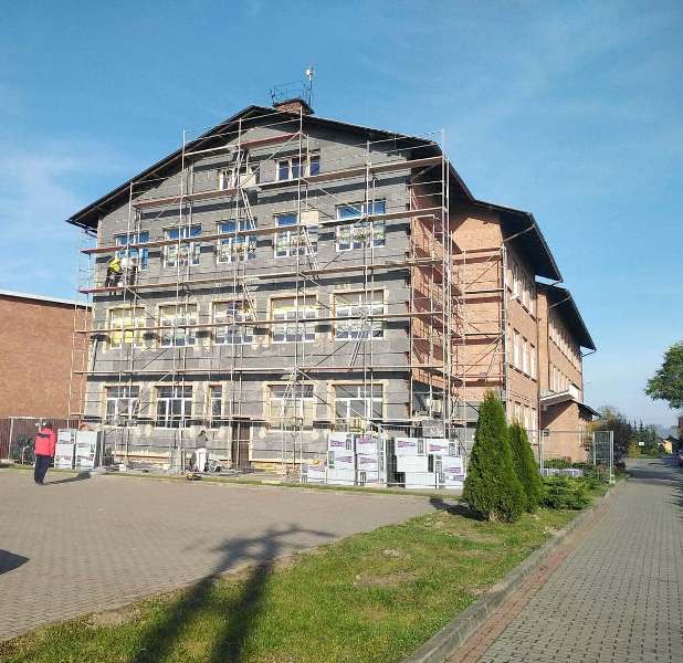 Trzykondygnacyjny budynek w trakcie remontu z rusztowaniem, ceglane ściany i liczne białe okna, na pierwszym planie chodnik i trawnik z krzewami, jasne niebo.