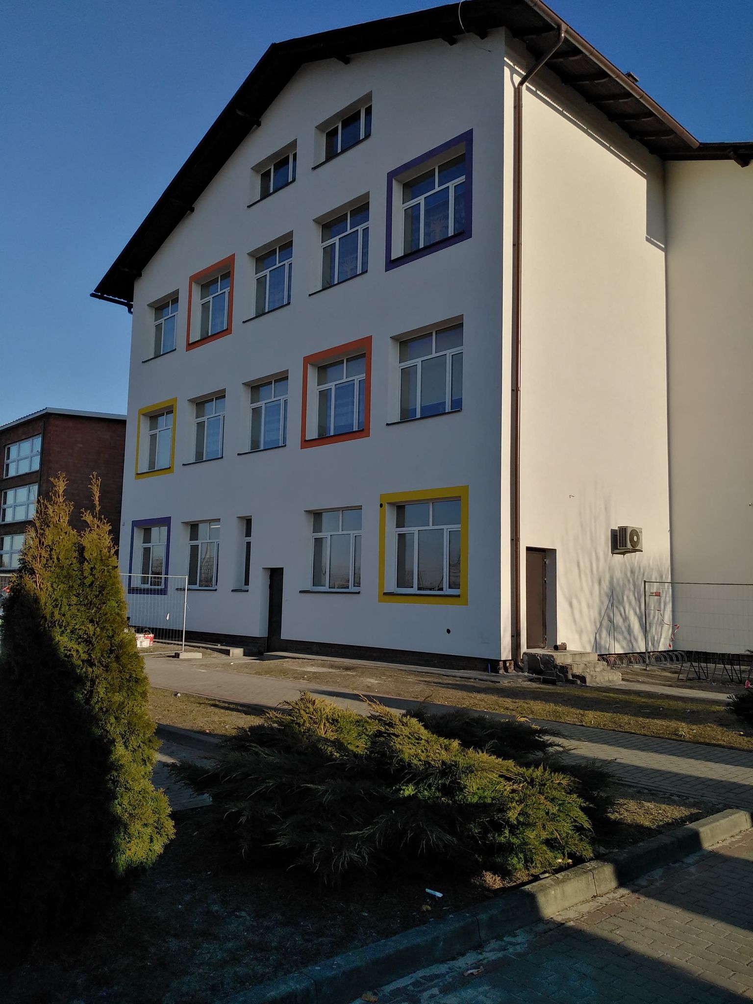 Biały budynek z trzema kondygnacjami i dachem dwuspadowym, okna w ramkach pomarańczowych, żółtych i fioletowych, na pierwszym planie krzewy i chodnik.