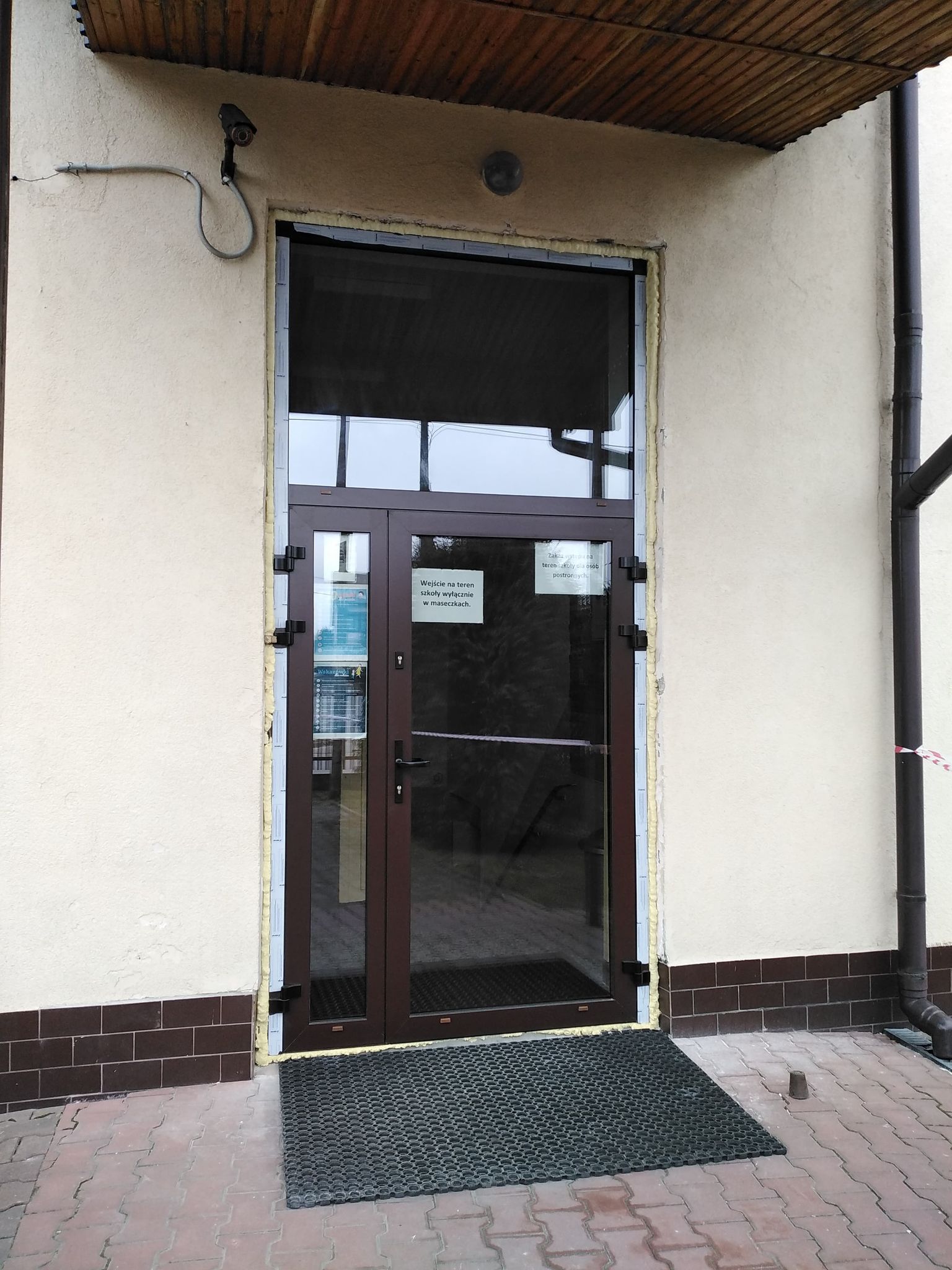 Szklane drzwi wejściowe z brązową ramą, nad nimi okno, po lewej kamera, po prawej rynna, pod drzwiami czarna gumowa wycieraczka, ściana kremowa z brązową cokołem.