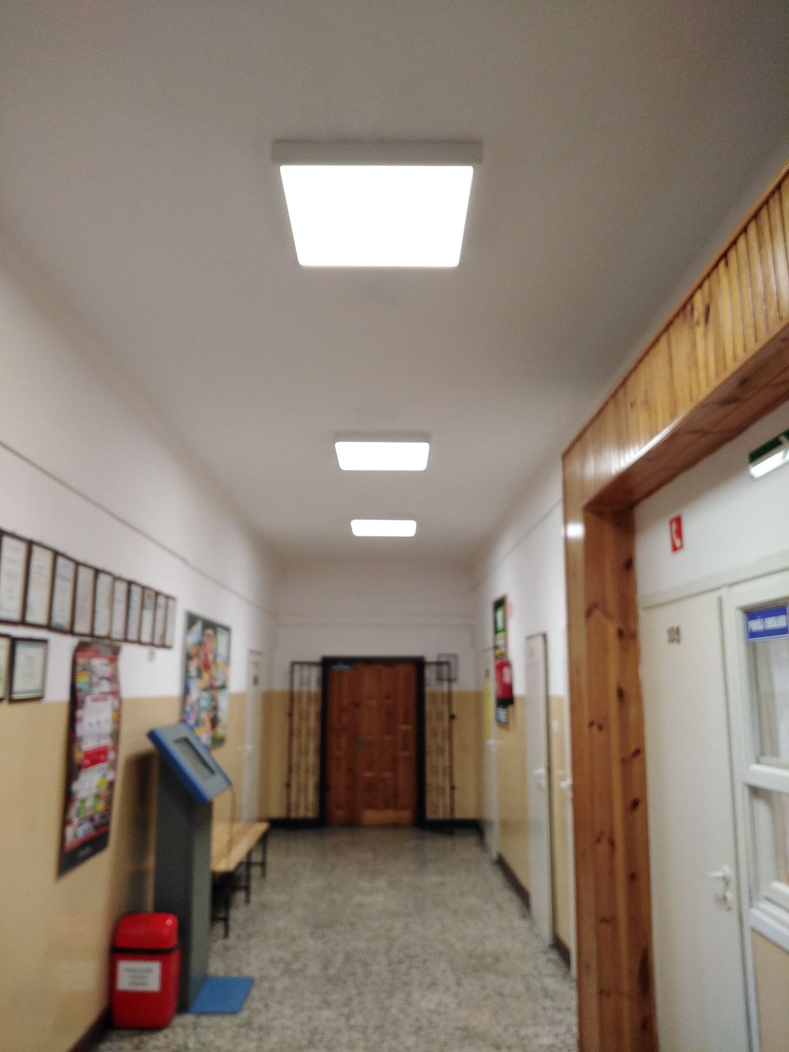 Korytarz z jasnymi sufitowymi lampami, ściany w dolnej części beżowe, w górnej białe, drewniana rama drzwi po prawej stronie, ławka i tablica informacyjna po lewej.