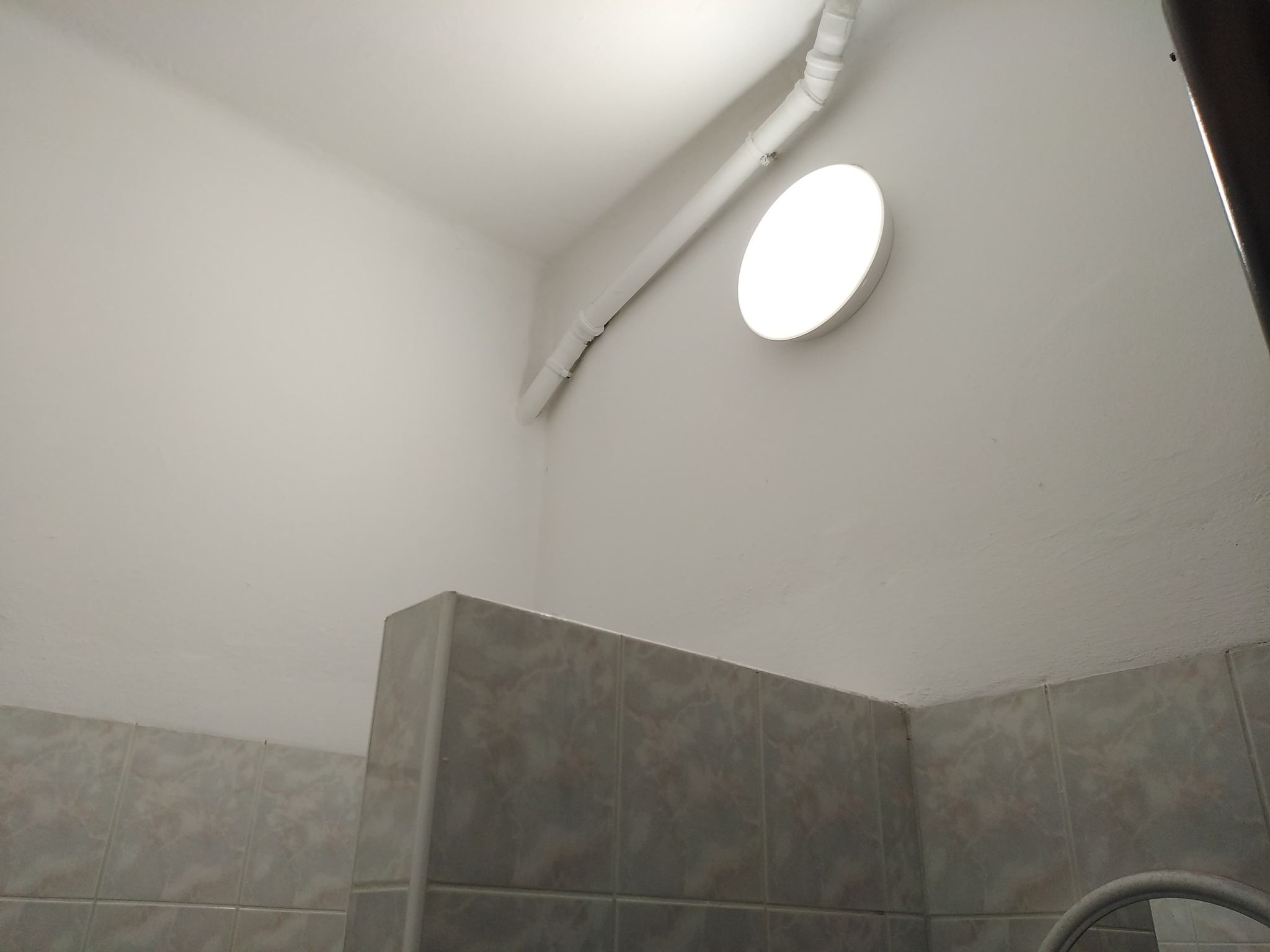 Białe ściany i sufit, szare płytki ceramiczne, biały okrągły plafon oraz biały plastikowy przewód instalacyjny na suficie.