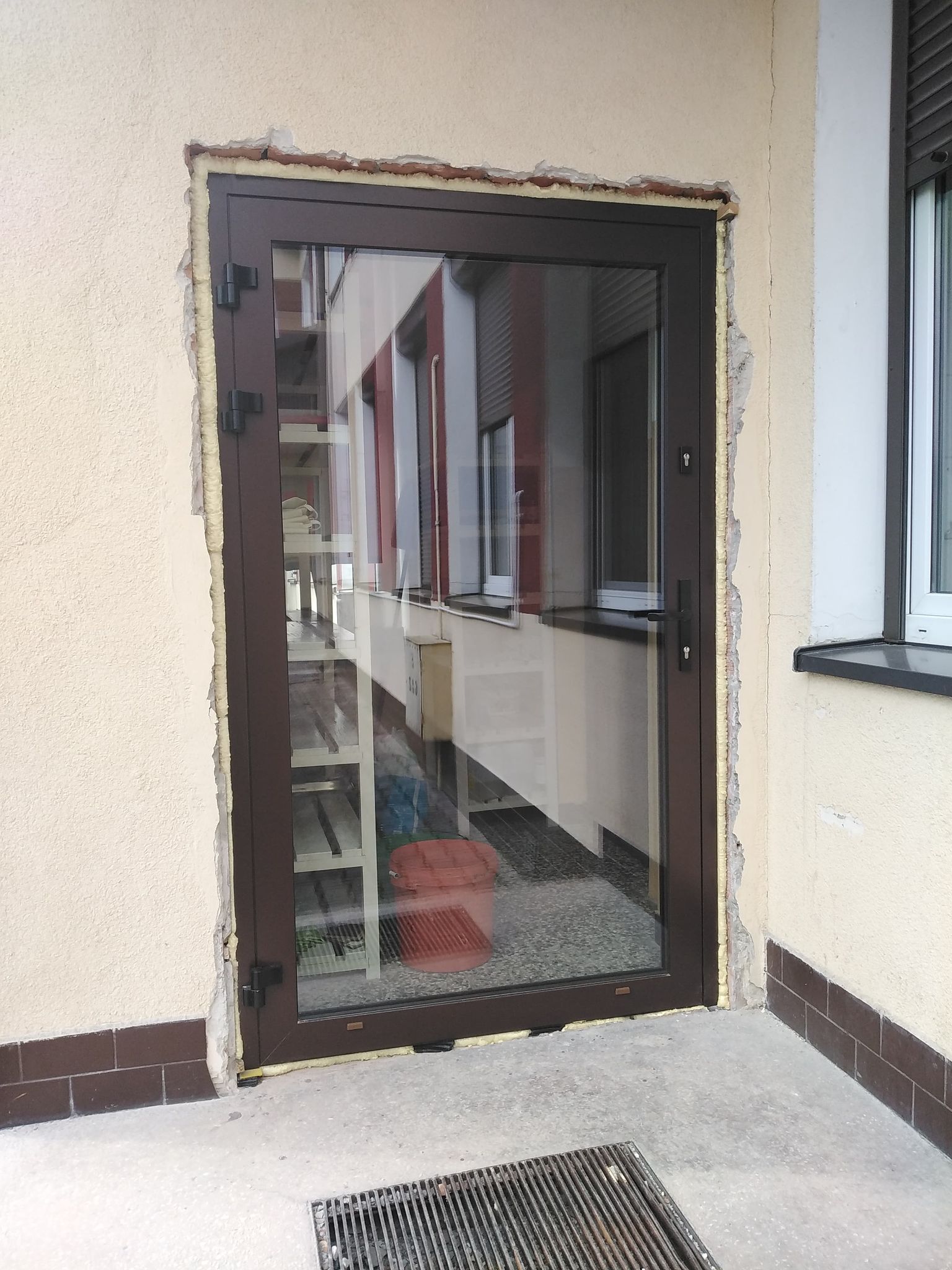 Ciemnobrązowe drzwi ze szkłem w ścianie budynku z odsłoniętą pianką montażową i cegłami, betonowa podłoga z metalową kratką.