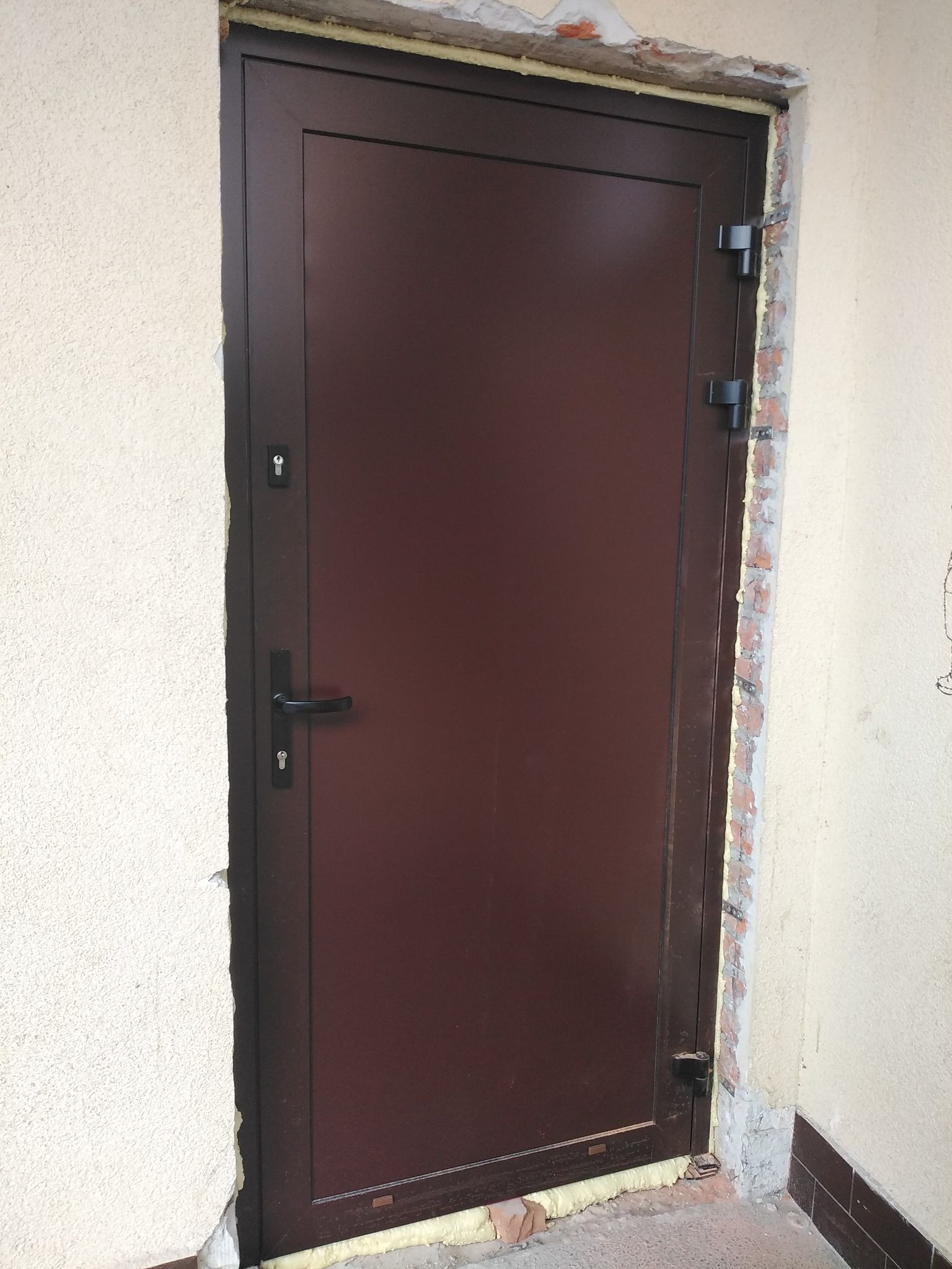 Brązowe, metalowe drzwi z czarną klamką osadzone w ścianie o jasnym tynku, z widoczną pianką montażową i odsłoniętą cegłą wokół ościeżnicy.