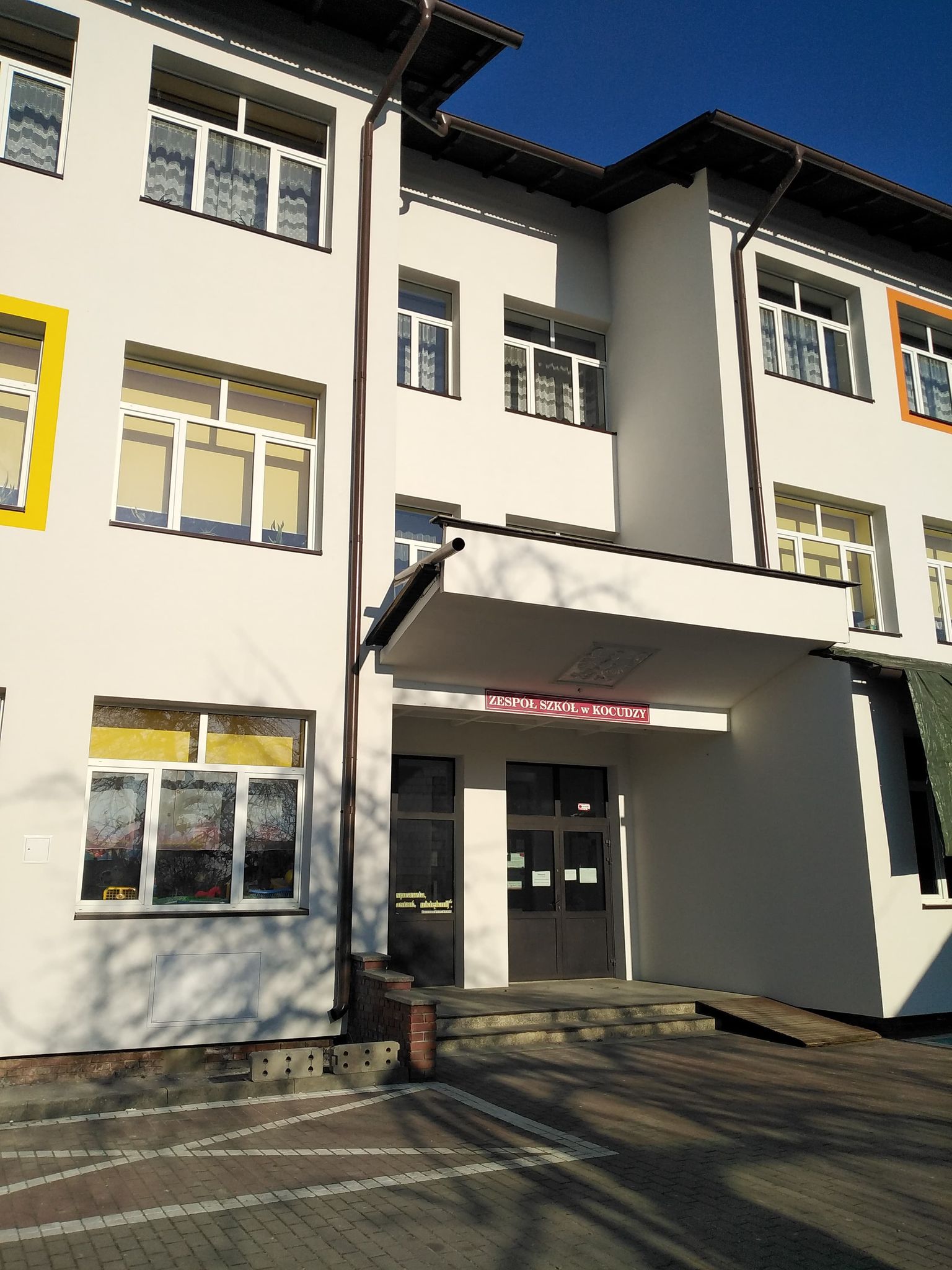 Biały budynek szkoły z trzema kondygnacjami, czarnymi drzwiami wejściowymi, kolorowymi ramami okien, zadaszeniem nad wejściem i chodnikiem z cegieł pod nim.