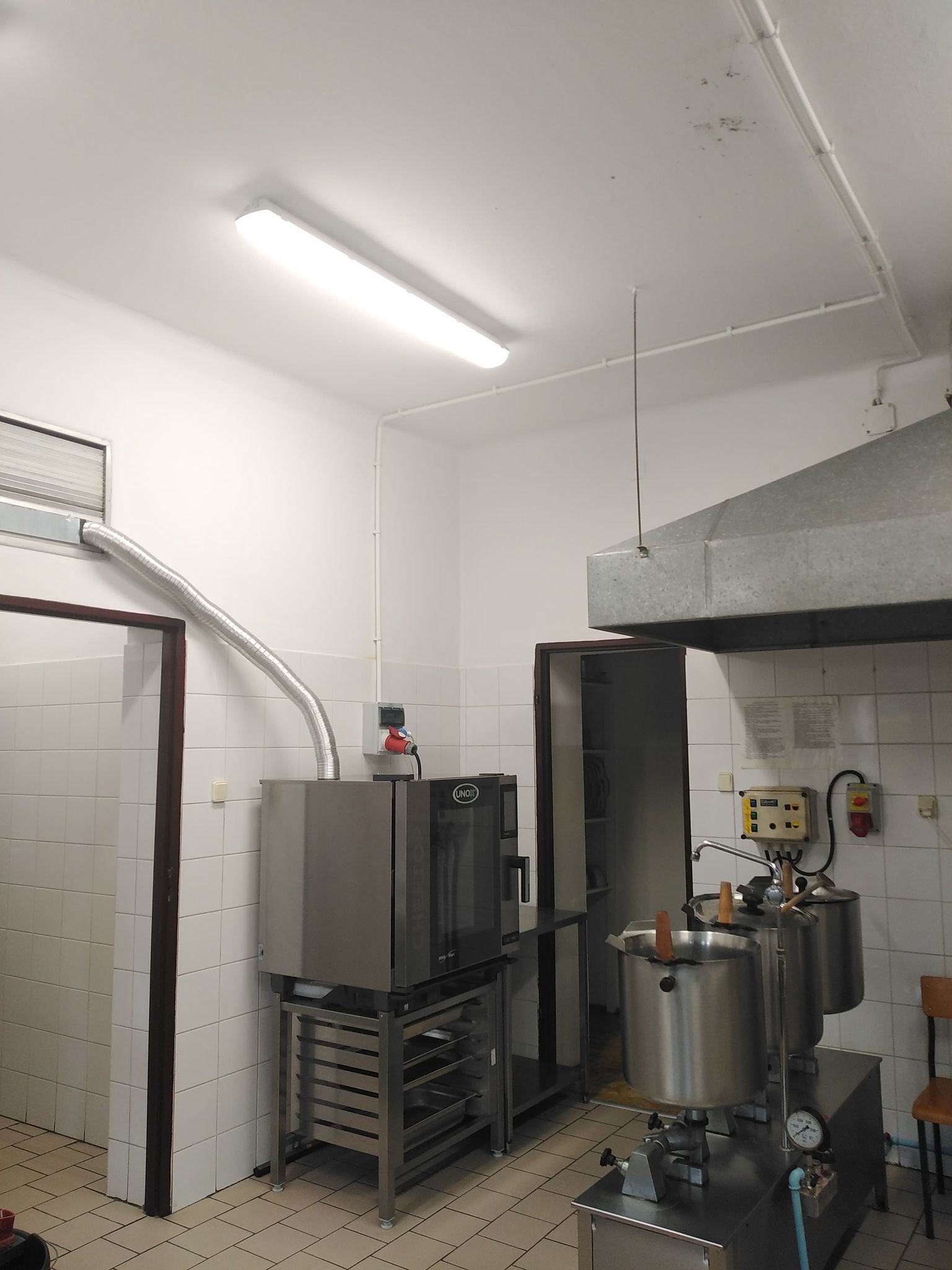 Biała kuchnia z szarym piekarnikiem na metalowej podstawie, dwoma dużymi metalowymi garnkami na palnikach, białe kafelki na ścianach, sufit z lampą i metalowym okapem.