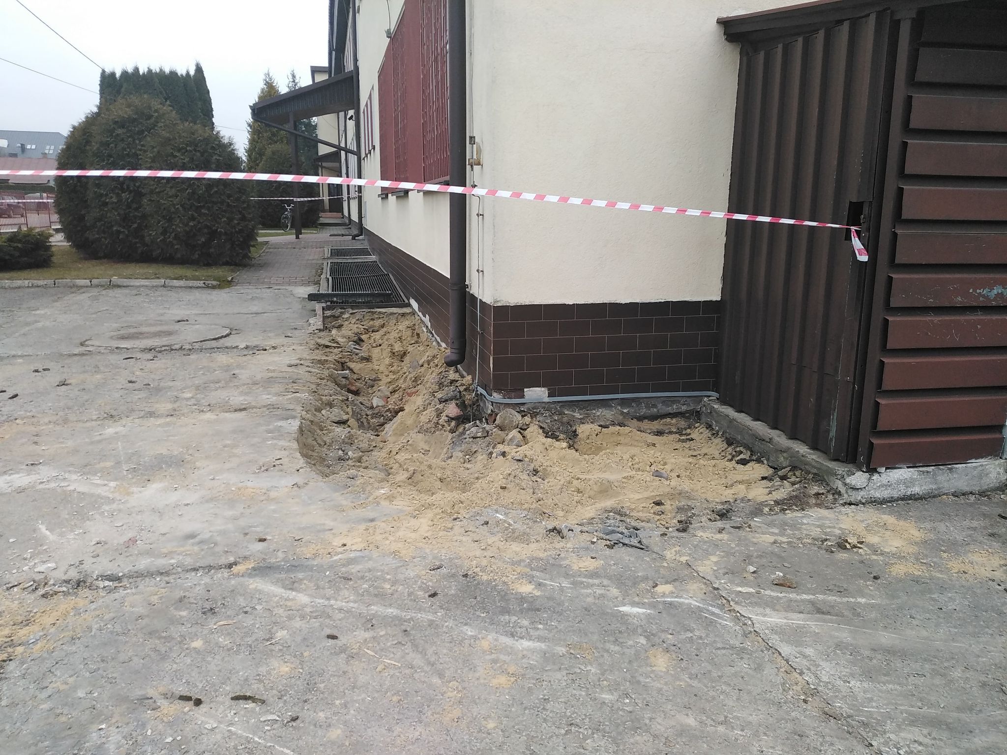 Fragment budynku z cokołem z brązowych płytek, wykop ziemny przy fundamentach, betonowa nawierzchnia, czerwona taśma ostrzegawcza rozciągnięta między budynkiem a metalową ścianą.