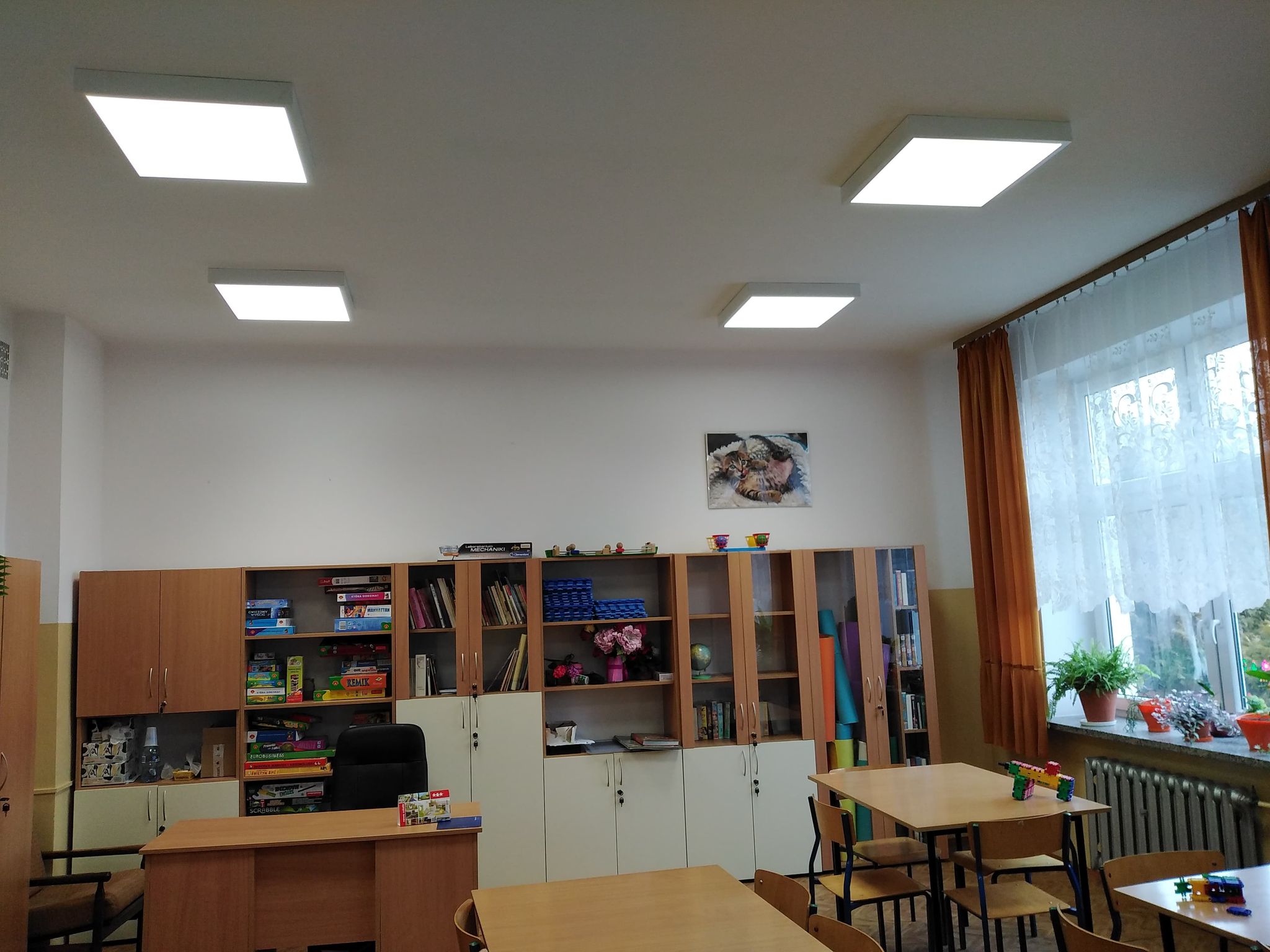 Klasyczna sala z jasnymi ścianami, drewniane biurko z czarnym fotelem, regały z półkami pełnymi książek, gier planszowych i dekoracji, duże okno z białą firaną i pomarańczowymi zasłonami, cztery kwadratowe lampy sufitowe.
