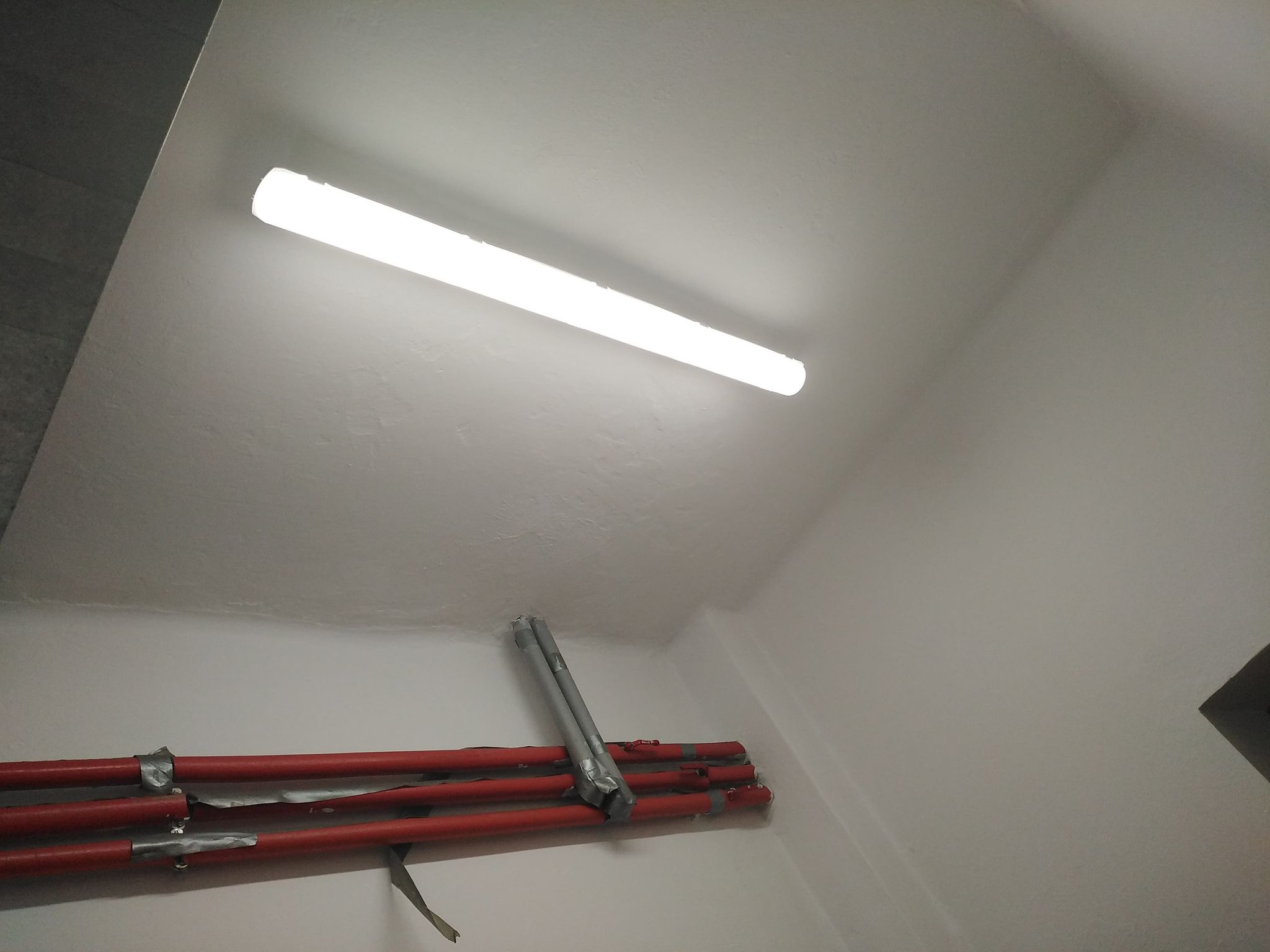 biały sufit z jasną świetlówką i czerwonymi rurami przymocowanymi do ściany, fragment szarej ściany po lewej stronie, biała ściana po prawej stronie