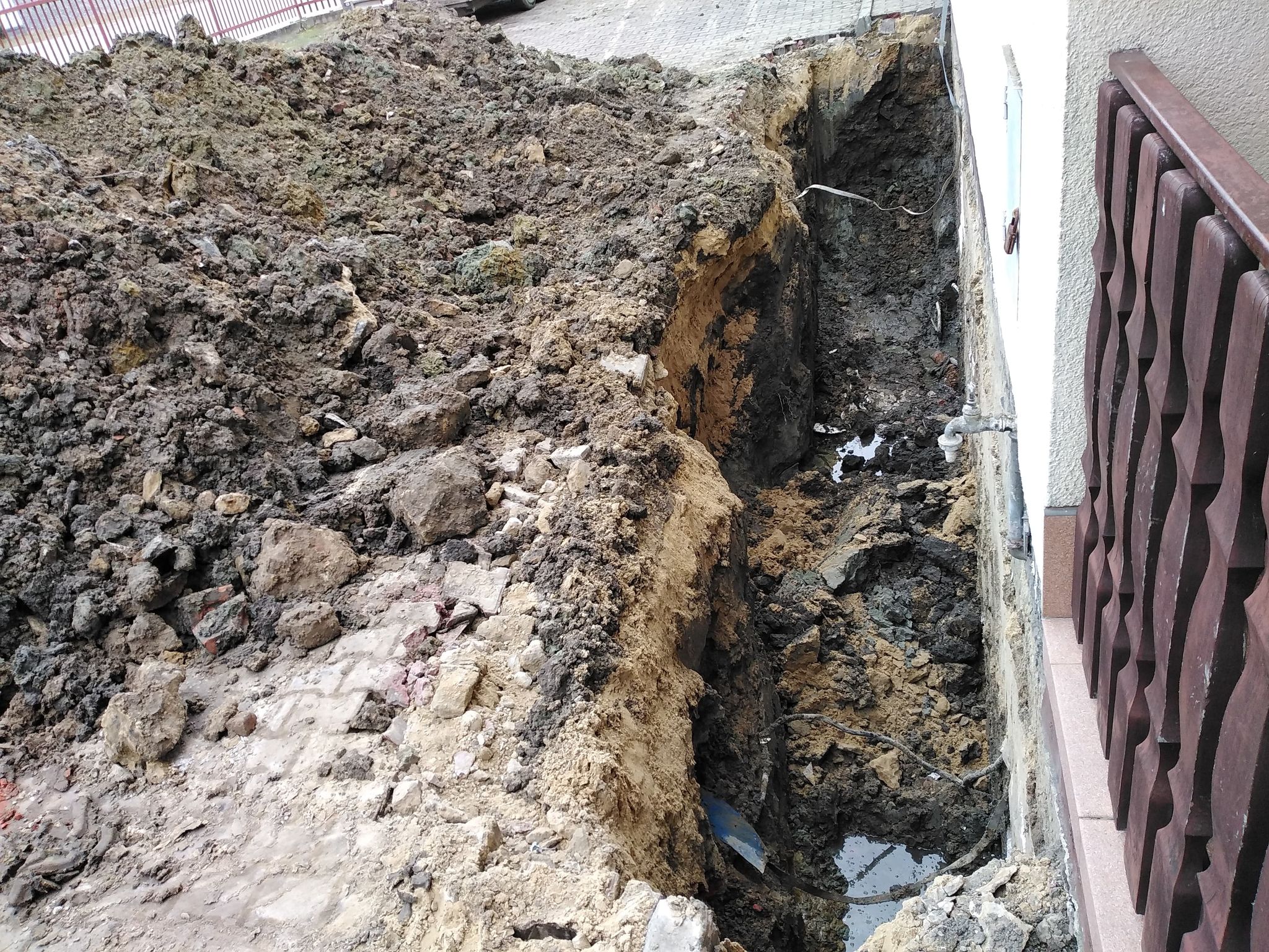 Wykopany rów obok budynku, ziemia o różnych odcieniach brązu, fragment płotu drewnianego i kran na ścianie budynku.