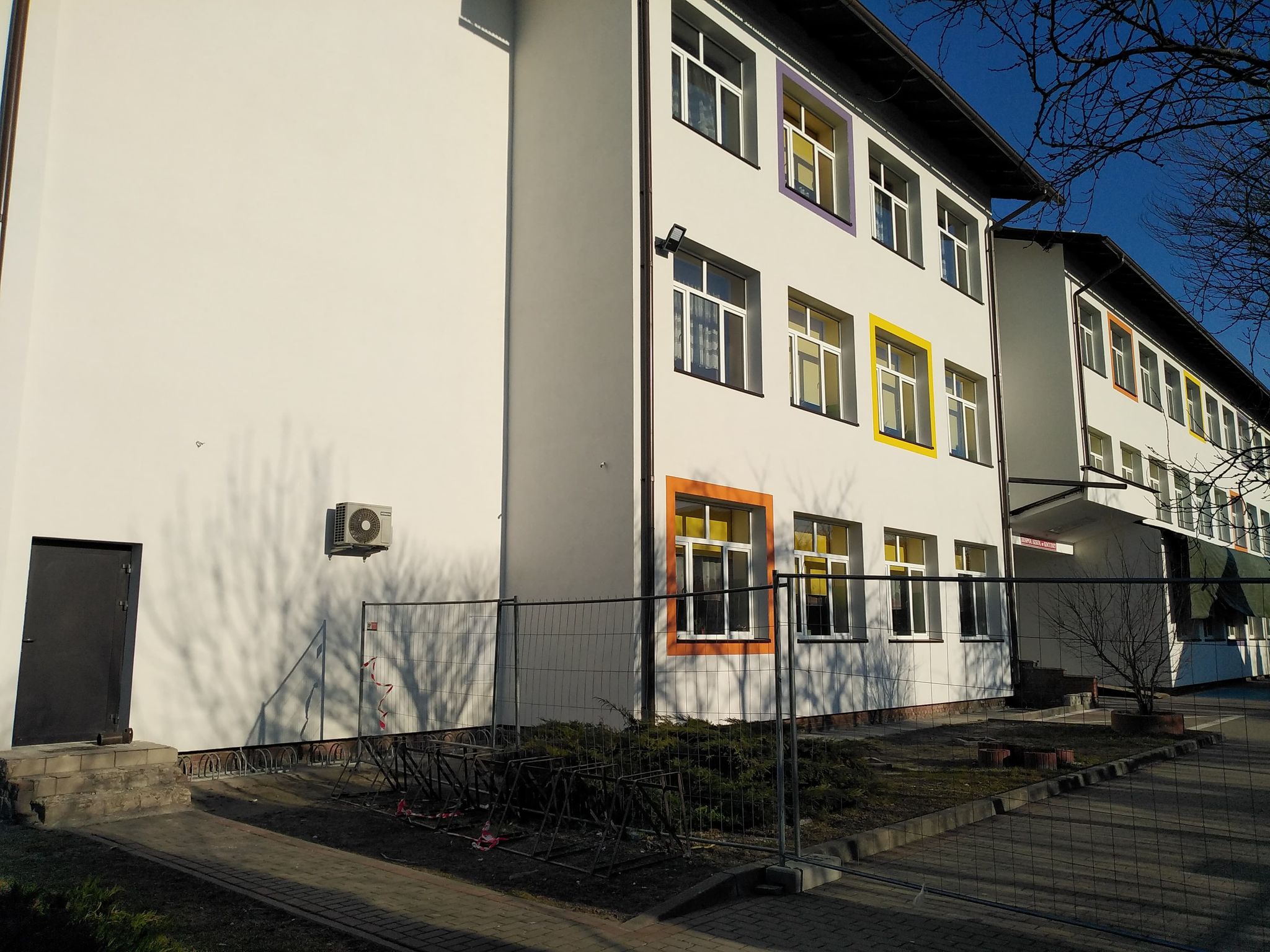 Biały budynek z wieloma oknami o kolorowych ramach, ogrodzenie metalowe, drzwi czarne, cienie drzew na ścianie, niebieskie niebo, chodnik z kostki.