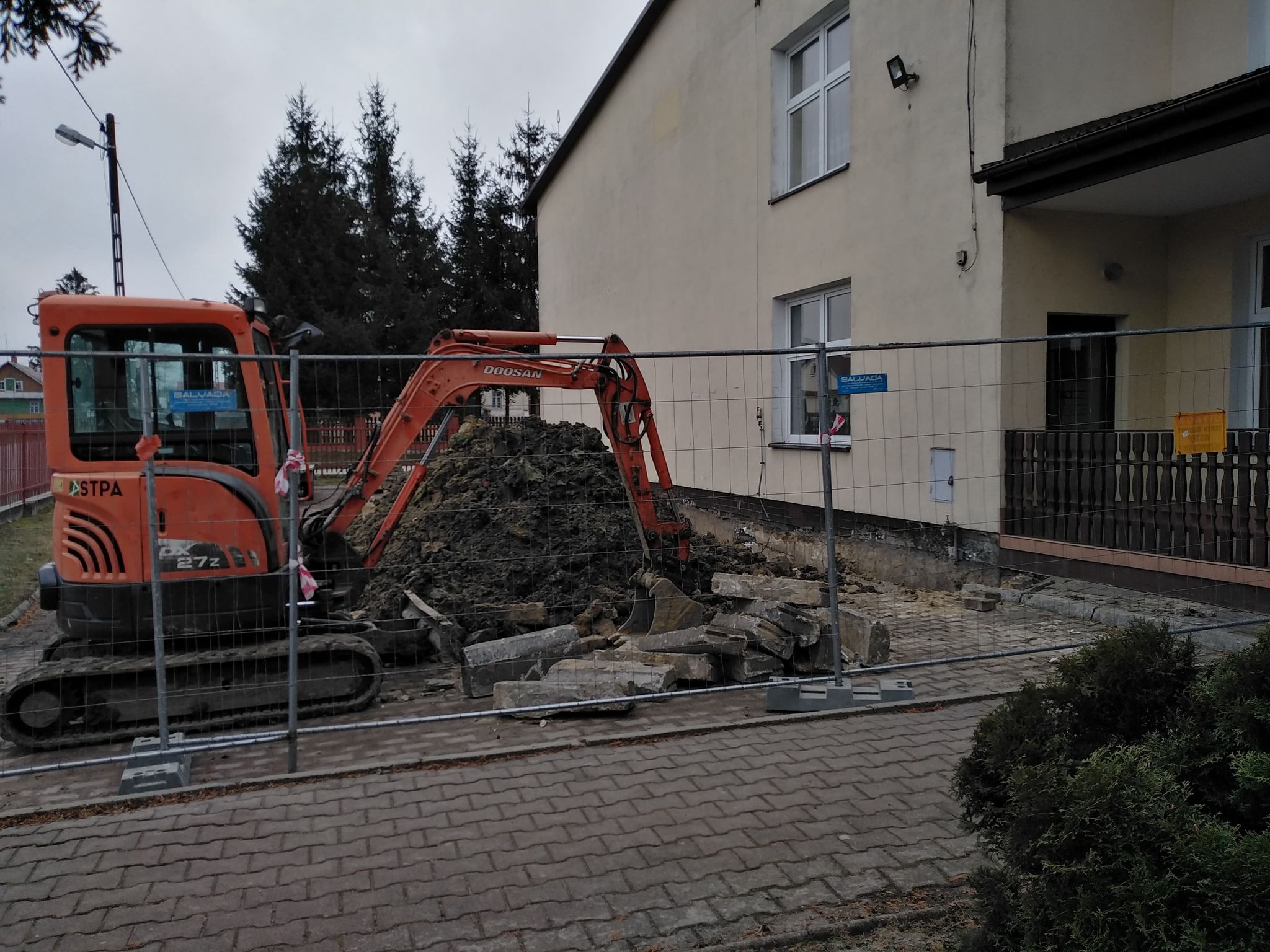 Pomarańczowa koparka na gąsienicach obok stosu ziemi i połamanych betonowych płyt, przed ogrodzeniem z siatki, przy szarym budynku z białymi oknami.