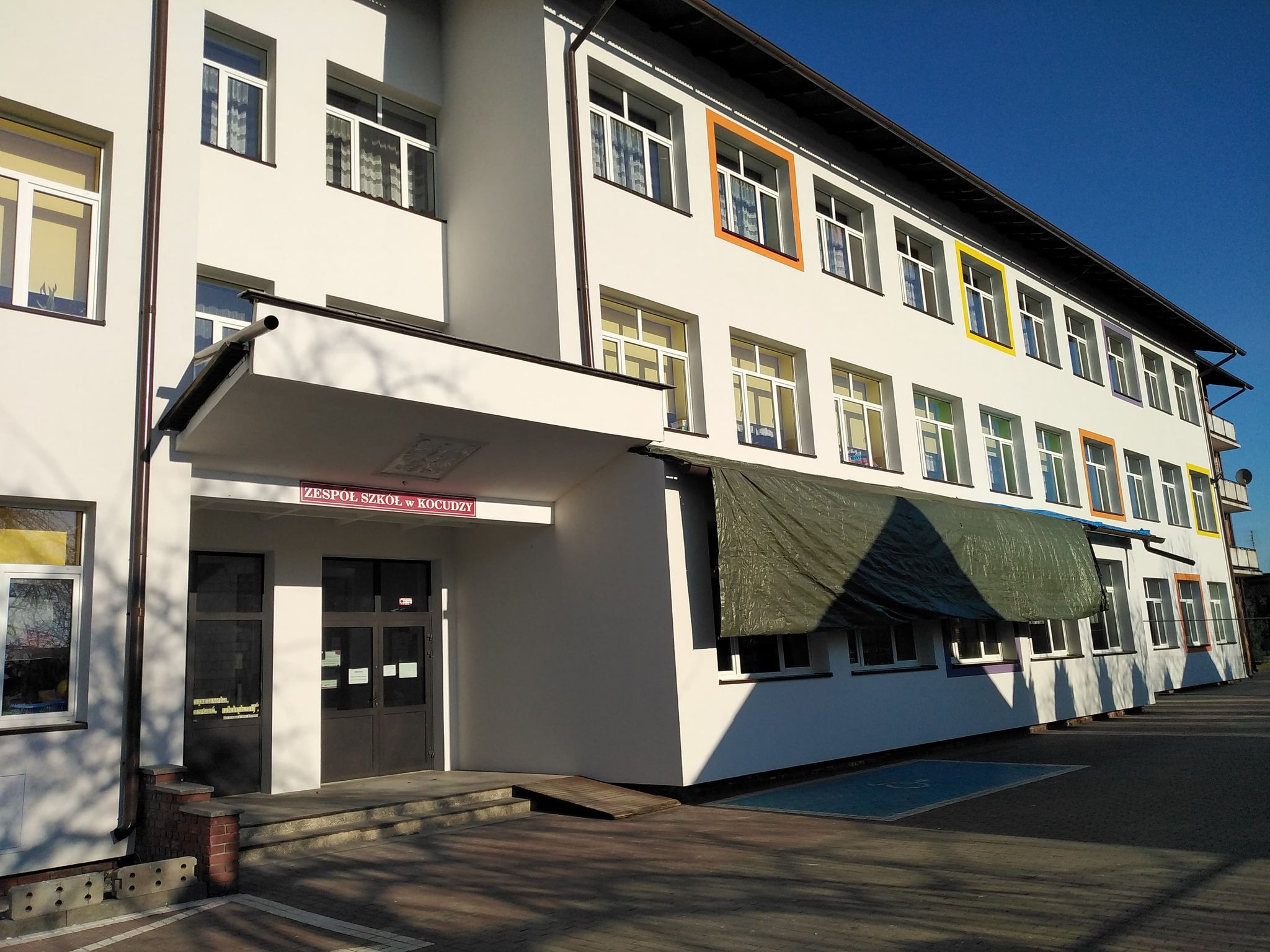 Biały budynek trzy piętrowy z wieloma oknami, niektóre obramowane na żółto, pomarańczowo i fioletowo, szare drzwi wejściowe, zielony brezent nad oknami, niebieskie niebo.