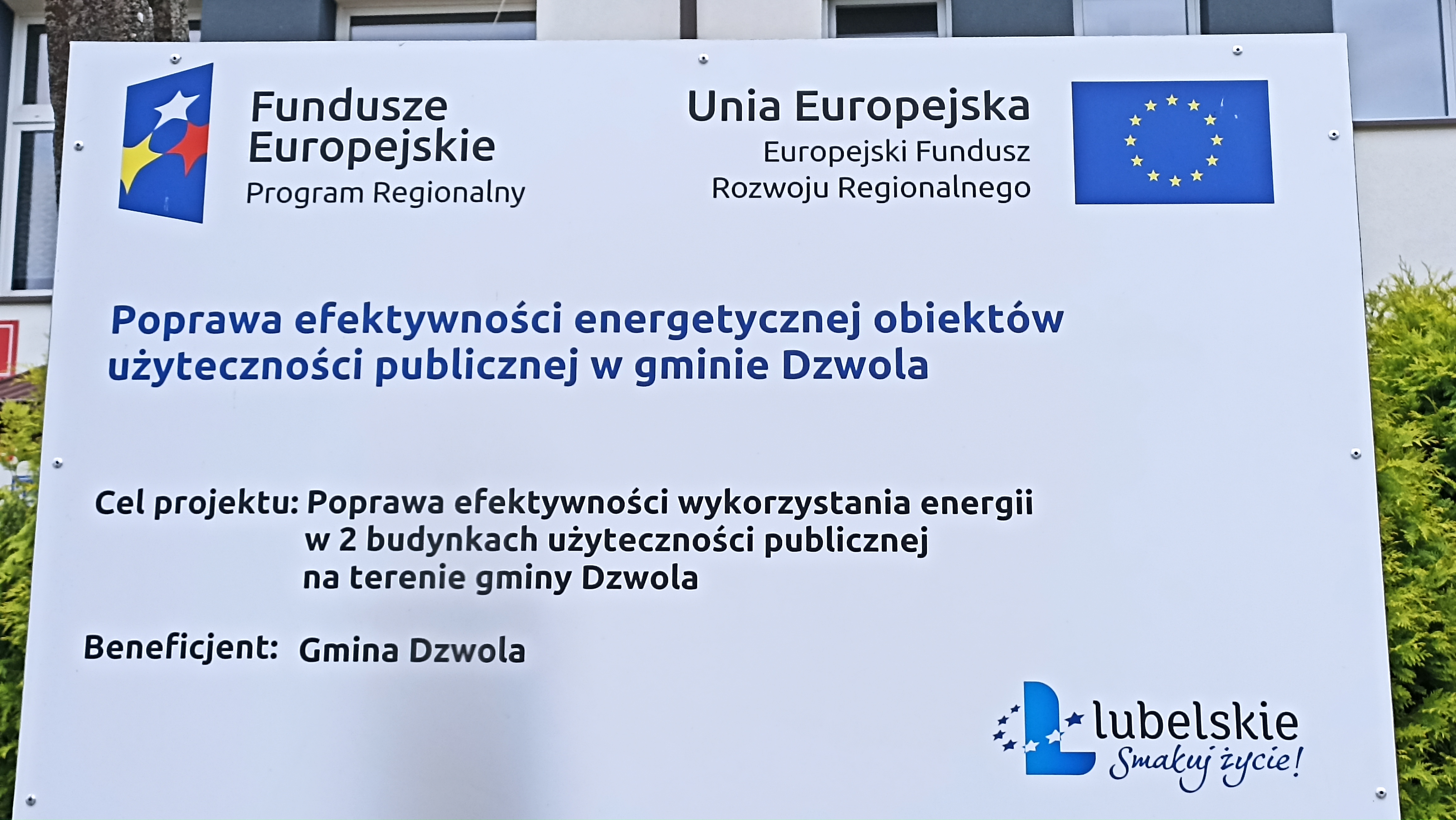 Tablica informacyjna z napisem o poprawie efektywności energetycznej obiektów użyteczności publicznej w gminie Dzwola, logotypy Funduszy Europejskich i Unii Europejskiej, zielone krzewy w tle.