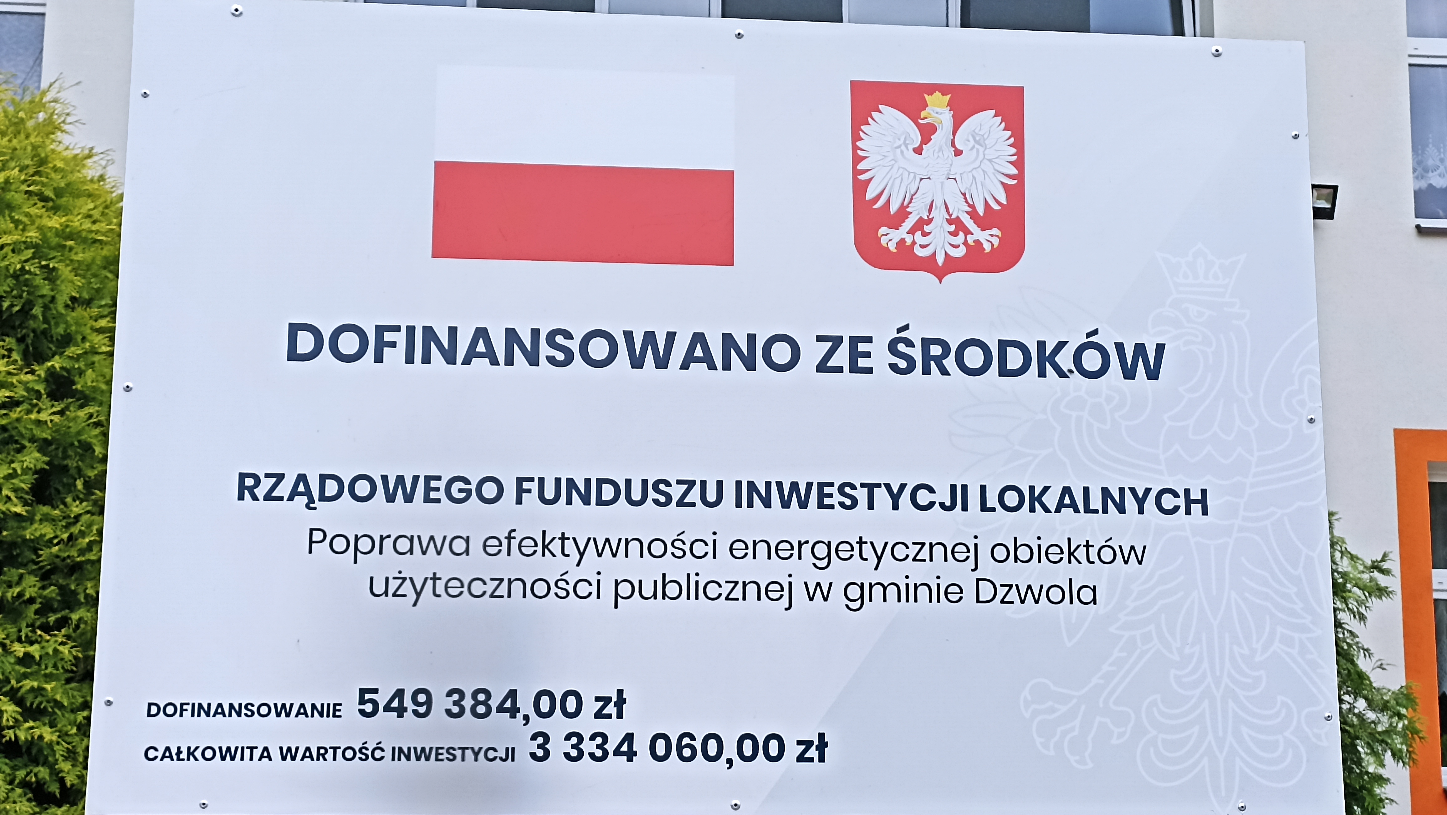 Tablica z polską flagą i godłem, informacja o dofinansowaniu rządowym, kwoty 549 384 zł i 3 334 060 zł, tło jasnoszare z wizerunkiem orła.