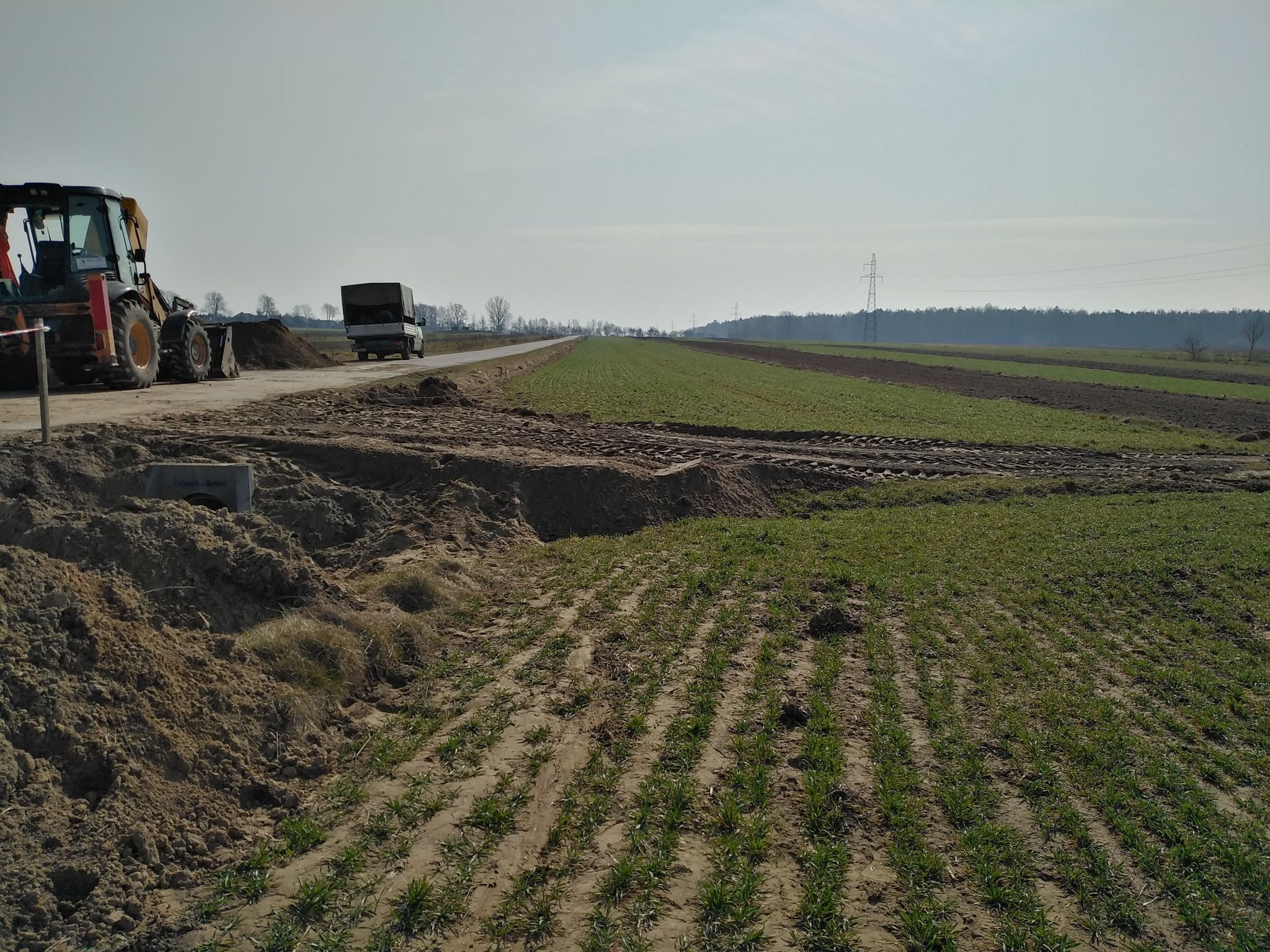 Pole z zielonymi młodymi roślinami, obok koparka i samochód ciężarowy na drodze, hałdy ziemi, linie energetyczne na tle nieba.