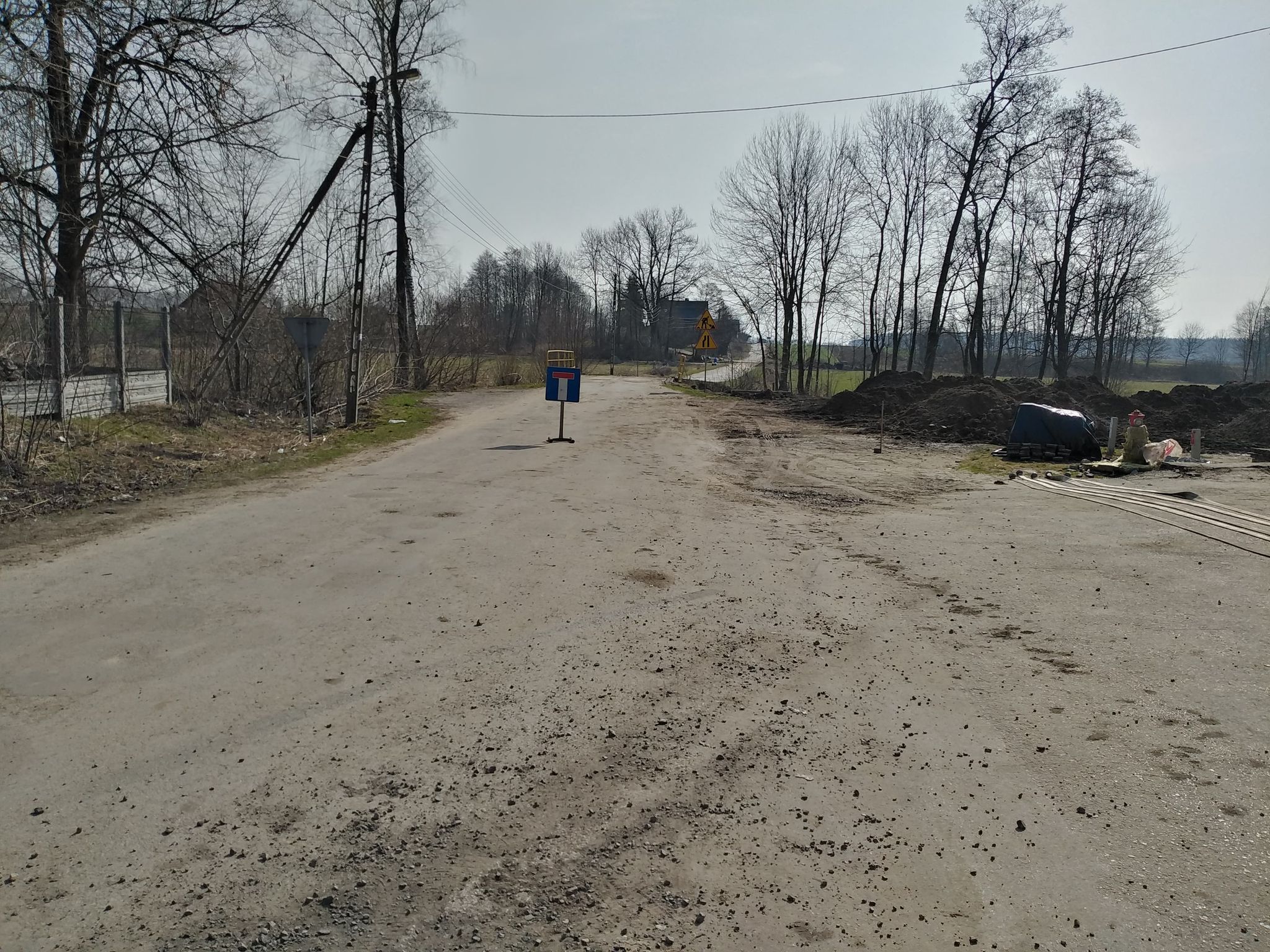 Droga z nawierzchnią asfaltową, po bokach nagie drzewa i znaki drogowe, po prawej hałdy ziemi i niebieski przykryty materiałem stos.