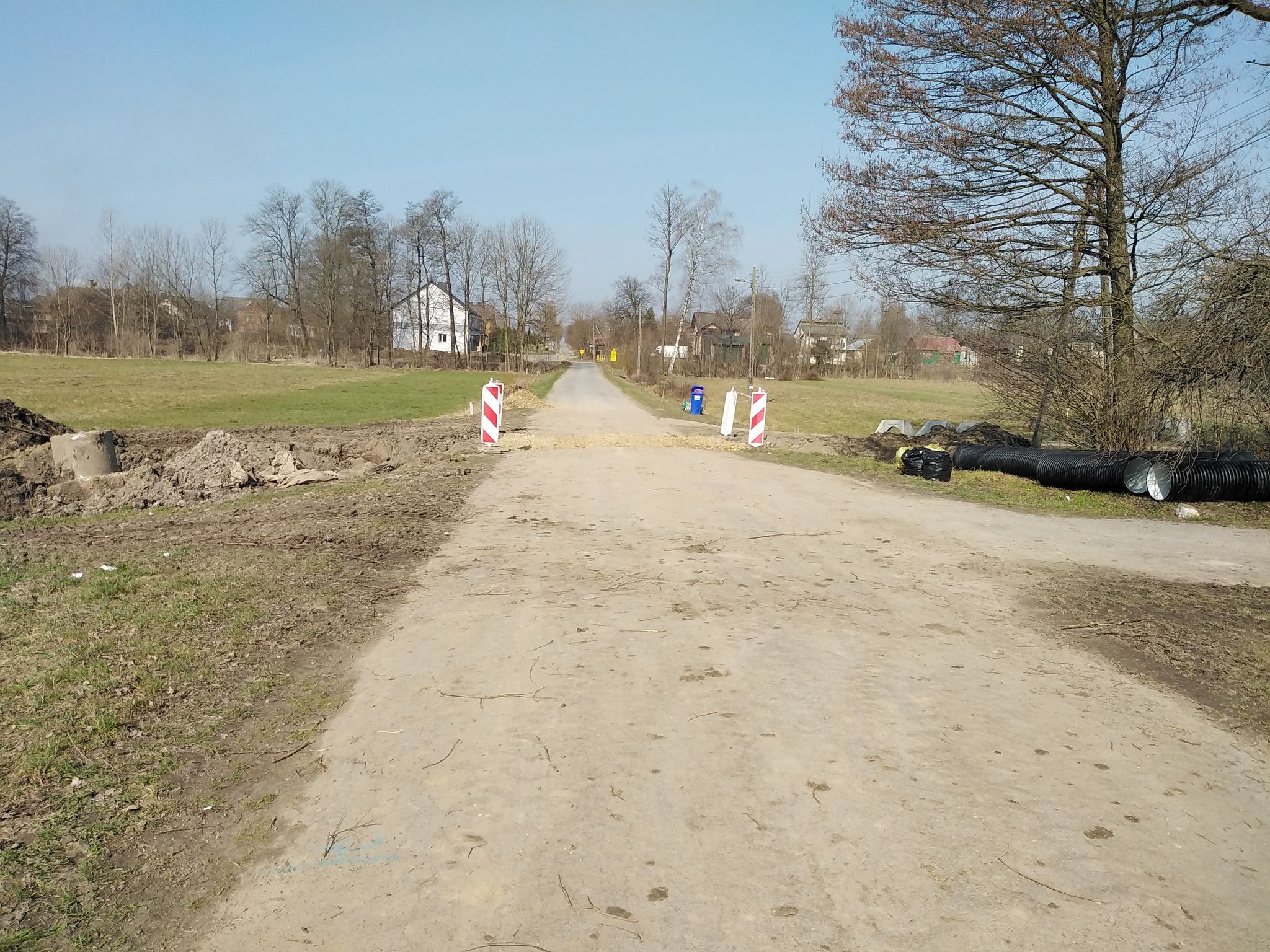Droga gruntowa z dwoma pionowymi znakami ostrzegawczymi, po bokach ziemia i trawa, w tle drzewa i domy, na niebie bezchmurne błękitne niebo.