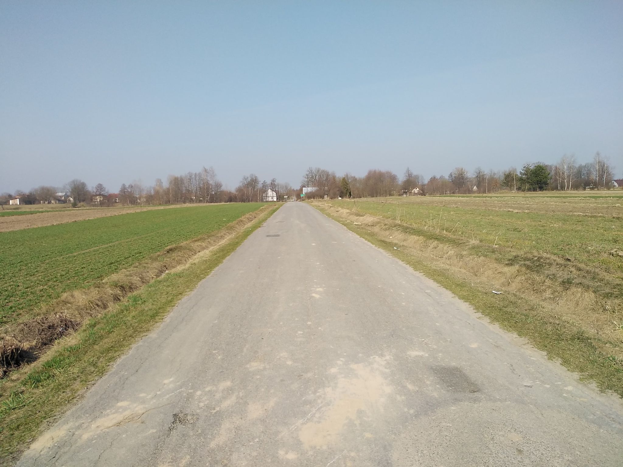 Wąska asfaltowa droga przez pola, po bokach zielone i brązowe łąki, w oddali drzewa i kilka domów pod jasnoniebieskim niebem.