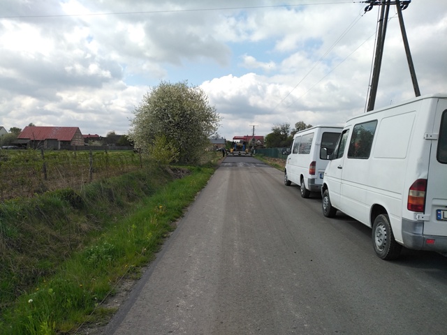 Wąska asfaltowa droga z trzema białymi dostawczymi samochodami zaparkowanymi po prawej stronie, z lewej trawa, krzewy i drzewa, w tle domy i pochmurne niebo.