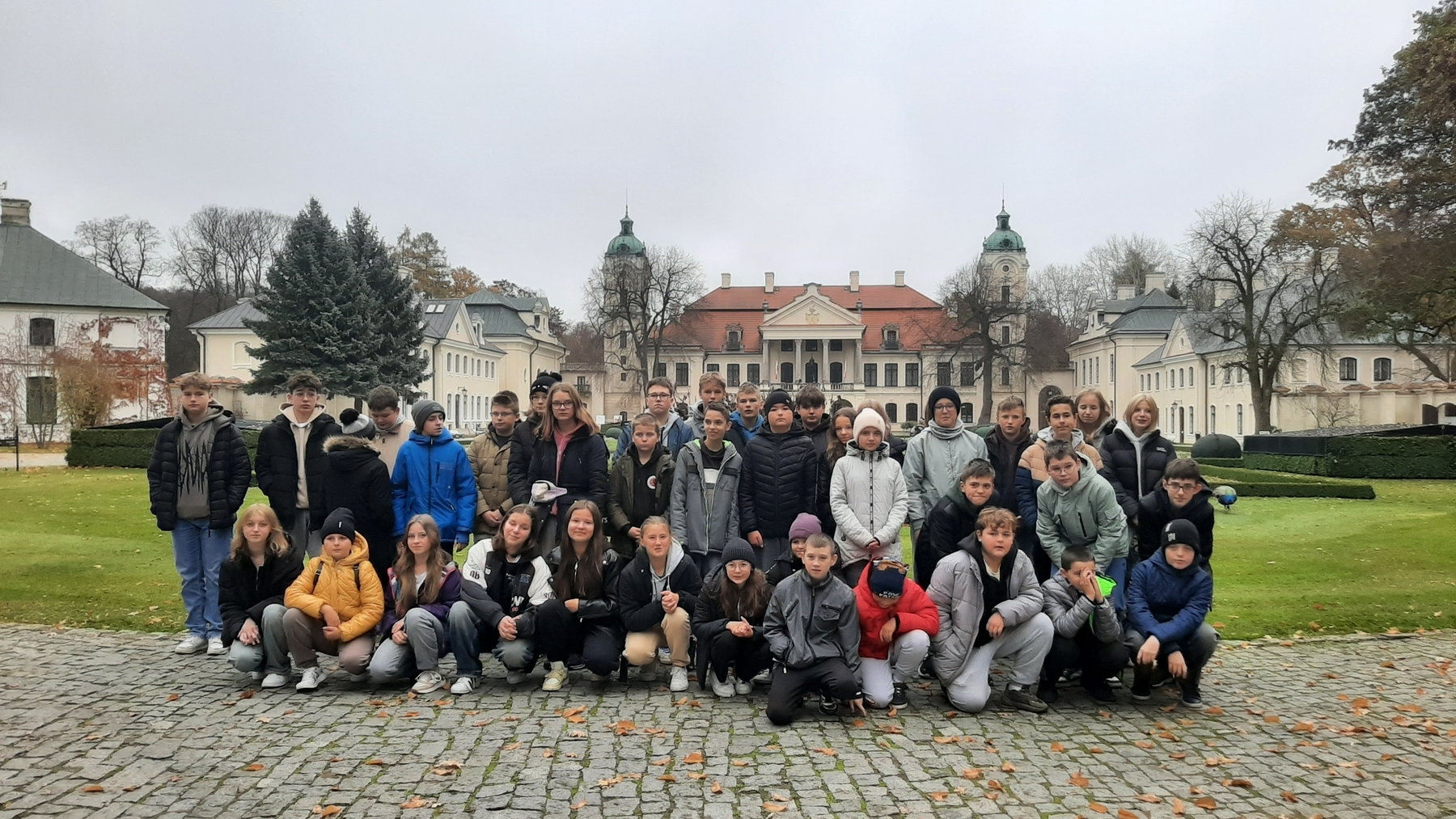 Grupa dzieci i nastolatków w kurtkach i czapkach na tle zabytkowego pałacu otoczonego zielonym trawnikiem i drzewami w pochmurny dzień.