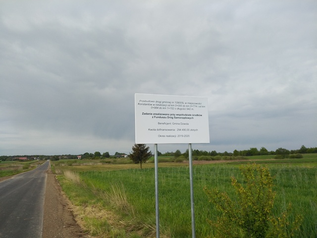 Tablica informacyjna z tekstem stojąca na metalowych słupach na tle zielonych pól i szarego nieba. Po lewej asfaltowa droga.