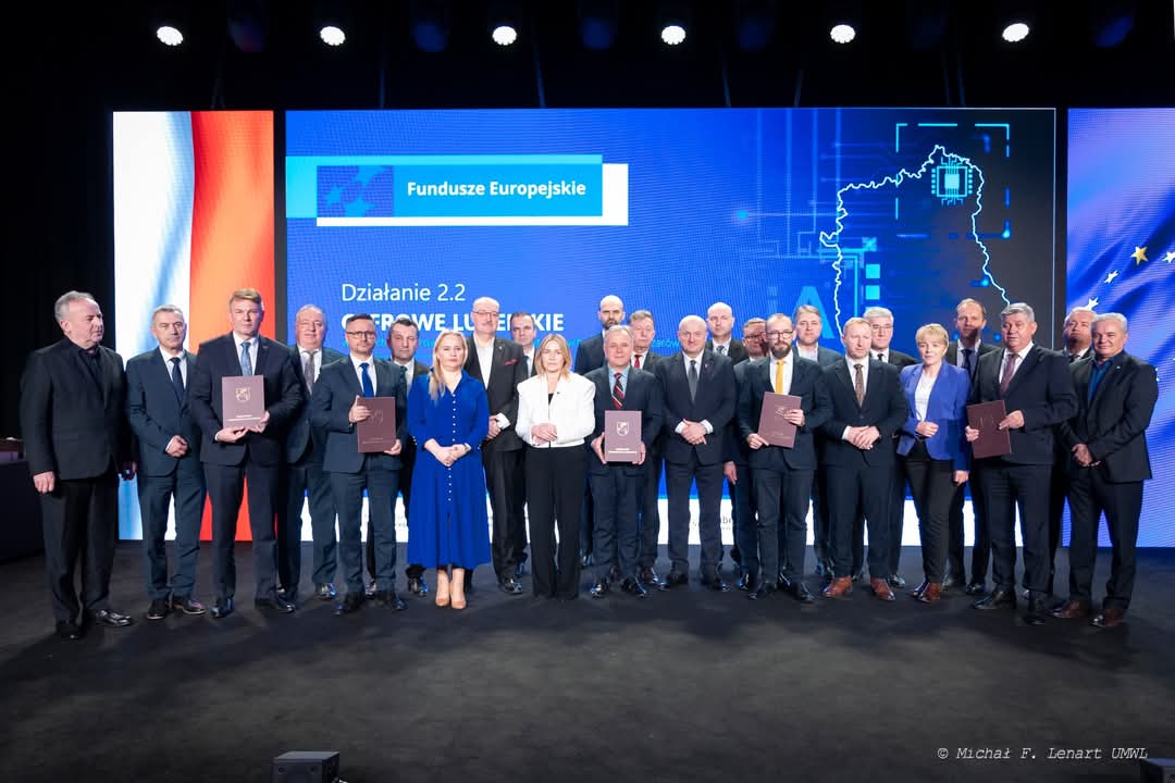 Grupa 21 osób w garniturach i sukienkach stoi na ciemnej scenie przed ekranem z niebieskim tłem, napisami i flagami Polski oraz Unii Europejskiej.