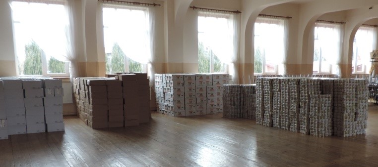 Wnętrze jasnego pomieszczenia z dużymi oknami i zasłonami, na drewnianej podłodze pięć dużych stosów kartonowych pudełek w różnych kolorach.