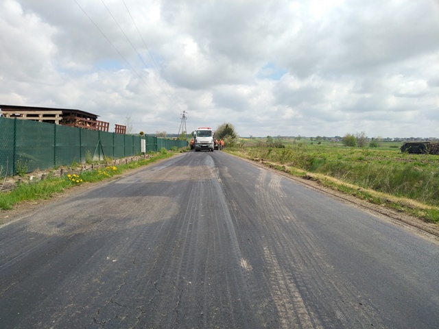 Asfaltowa droga z lekkimi śladami ścierania, pośrodku biały samochód dostawczy, po lewej zielony płot i budynek, po prawej pola uprawne pod pochmurnym niebem.
