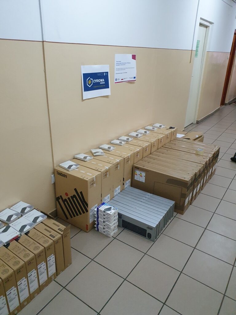 Rzędy kartonowych pudełek z elektroniką i akcesoriami na beżowej podłodze w korytarzu o jasnych ścianach, obok dwóch plakatów na ścianie.