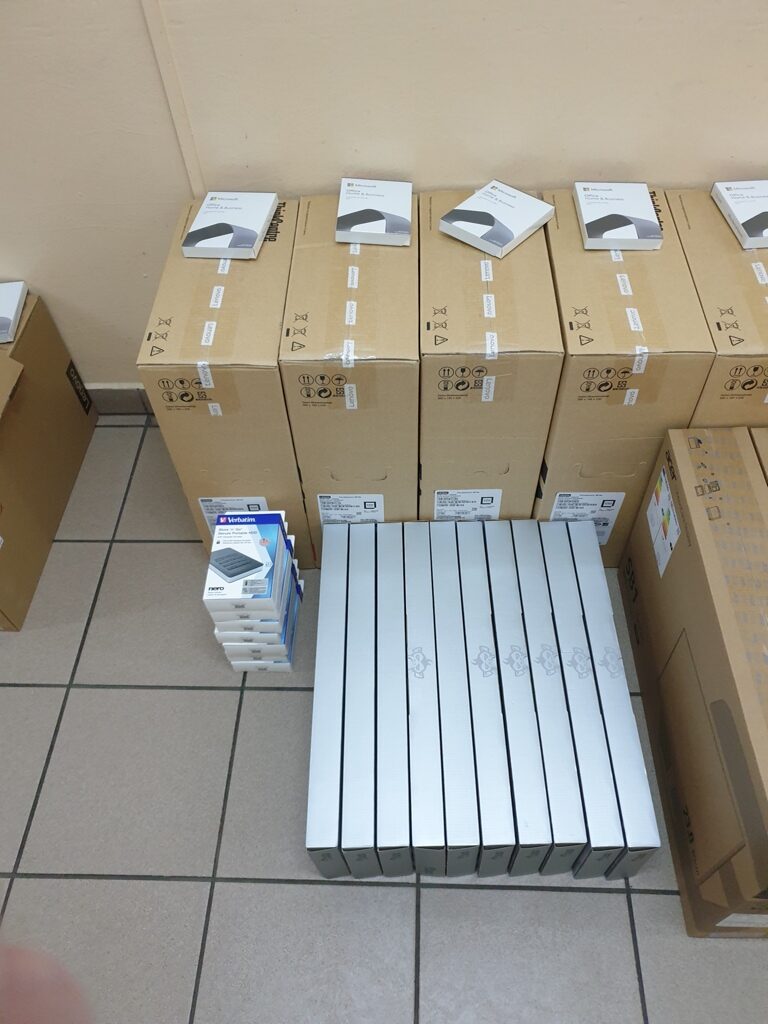 Stos kartonowych pudeł z etykietami, na podłodze jasnobrązowa płytka, na pudełkach białe opakowania z grafiką czarnego urządzenia i niebieskie opakowania mniejszych rozmiarów.