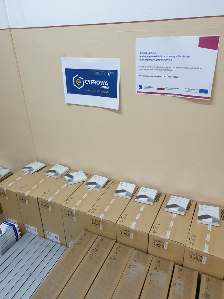 Stosy kartonowych pudełek na podłodze pod ścianą z dwoma plakatami, jeden z niebieskim tłem i napisem "Cyfrowa Gmina", drugi z tekstem informacyjnym na białym tle.