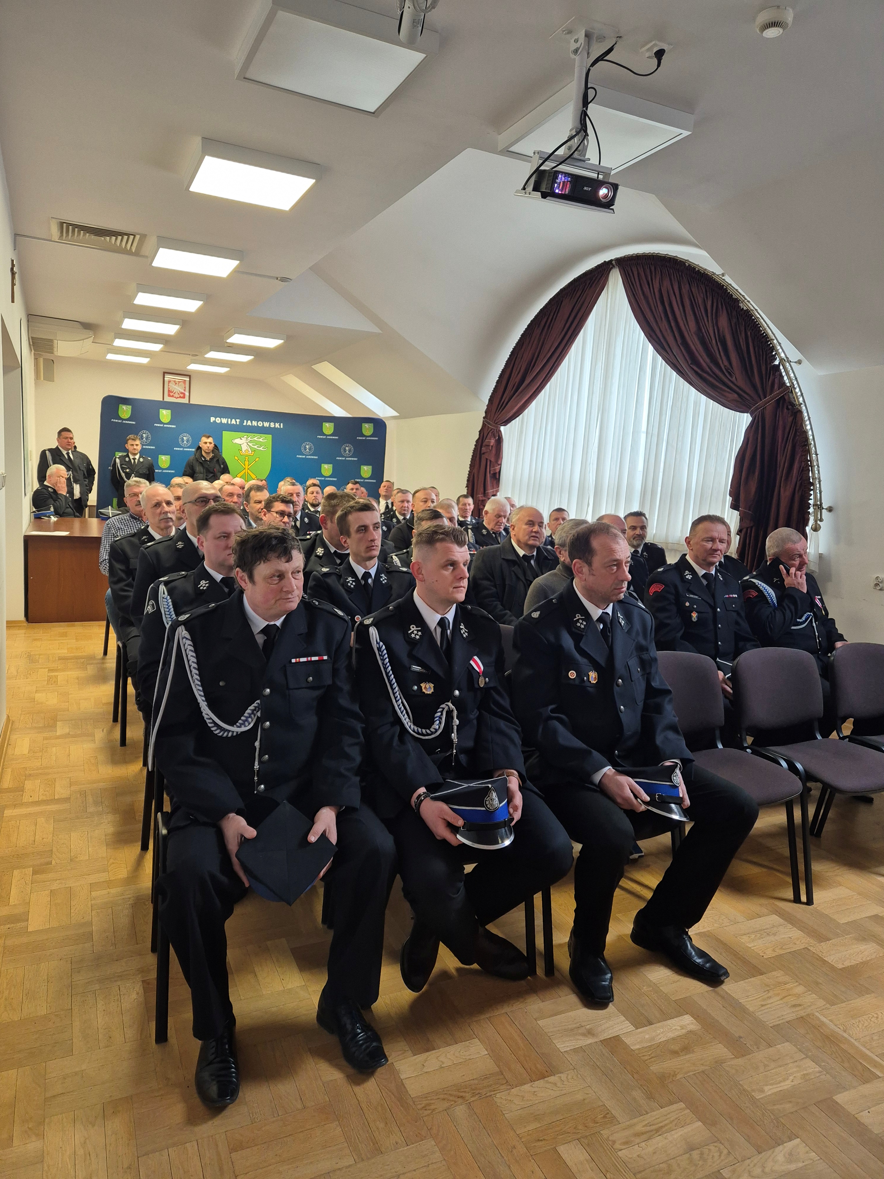 Grupa mężczyzn w mundurach siedzi na krzesłach w jasnym pomieszczeniu z drewnianą podłogą, dużym okrągłym oknem z bordowymi zasłonami i projektorem sufitowym.