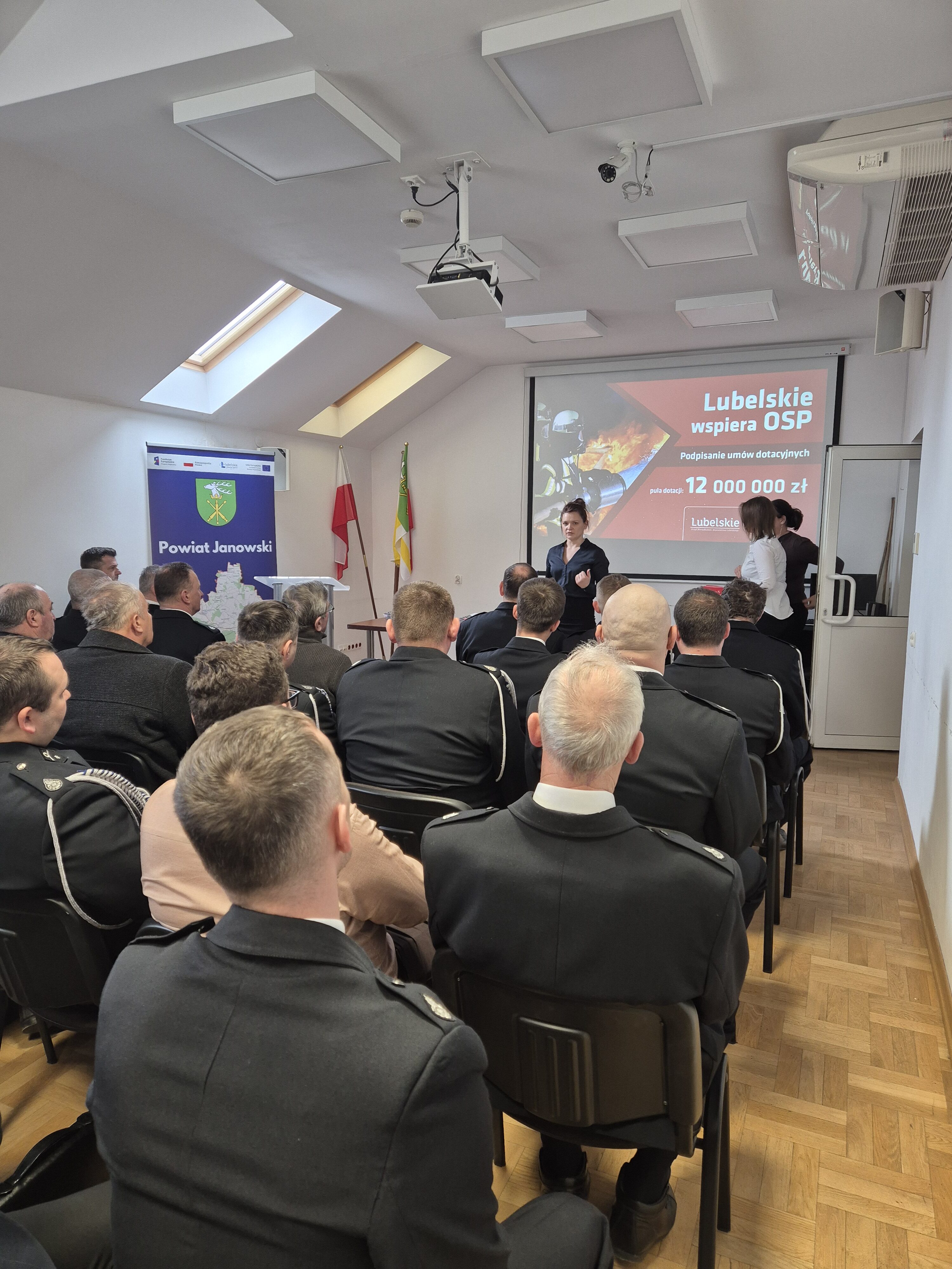 Grupa mężczyzn i kobiet w mundurach siedzi na krzesłach w sali konferencyjnej, na ścianie ekran z projekcją dotyczącą wsparcia OSP w województwie lubelskim.