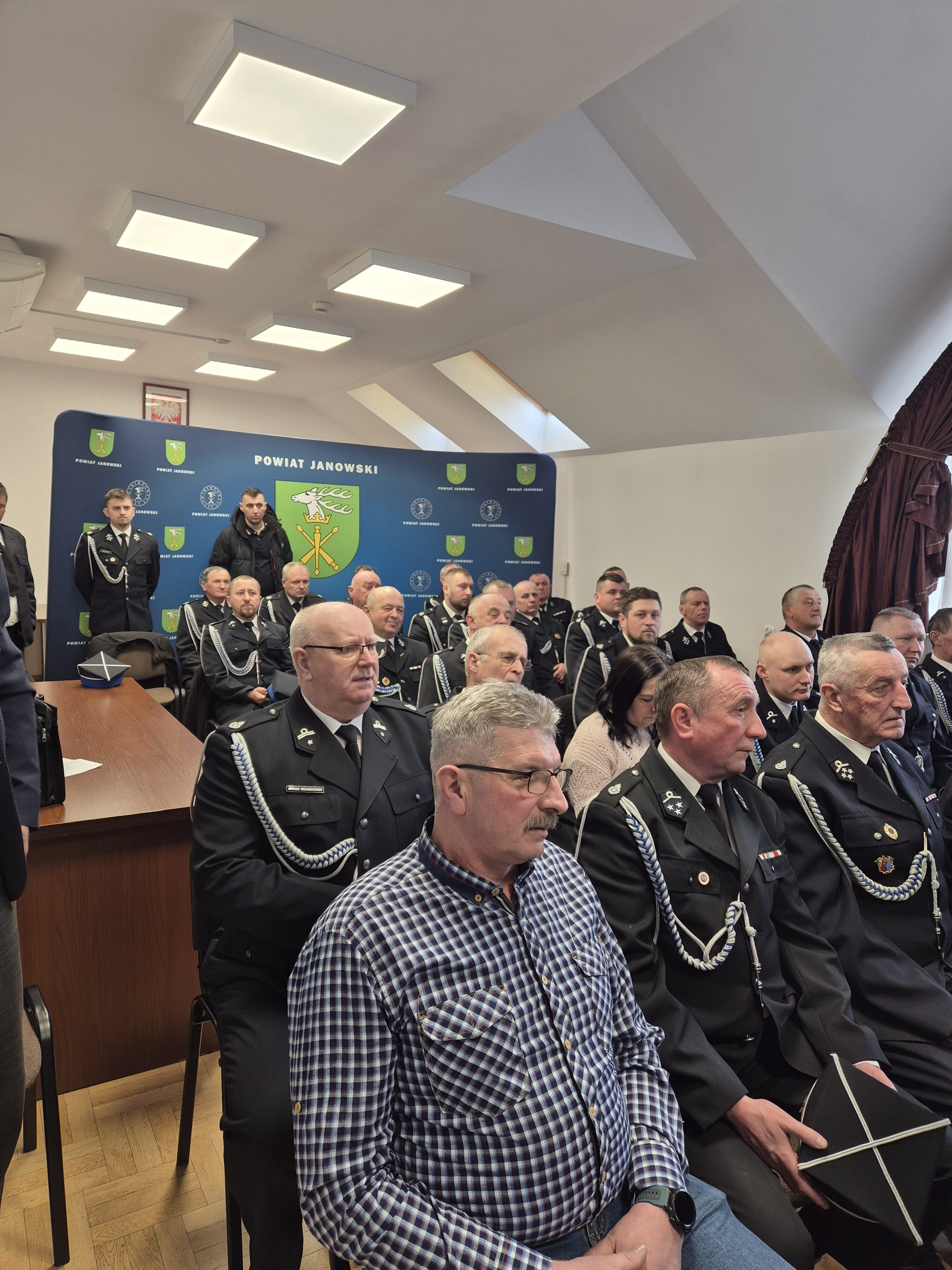 Grupa mężczyzn i jedna kobieta siedzący w sali, większość mężczyzn w mundurach strażackich, w tle niebieski baner z herbem i napisem "Powiat Janowski".