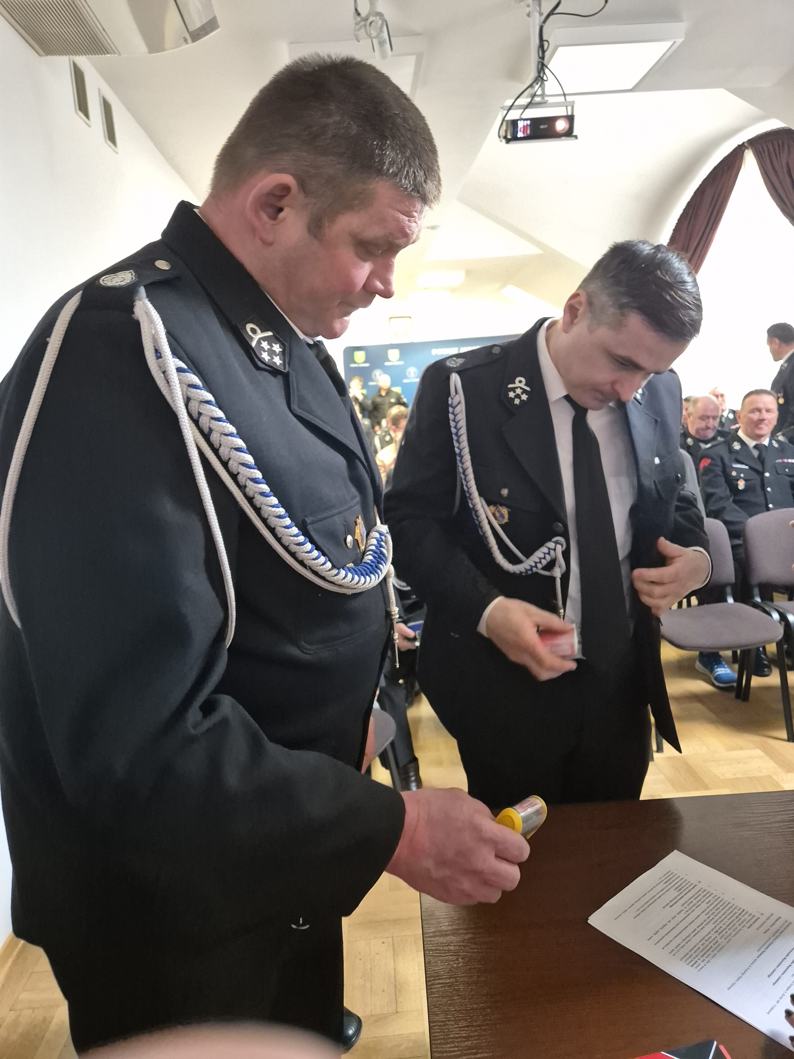 Dwóch mężczyzn w czarnych mundurach z białymi sznurami stoi przy stole z dokumentem, w tle inne osoby siedzące na krzesłach w jasnym pomieszczeniu.