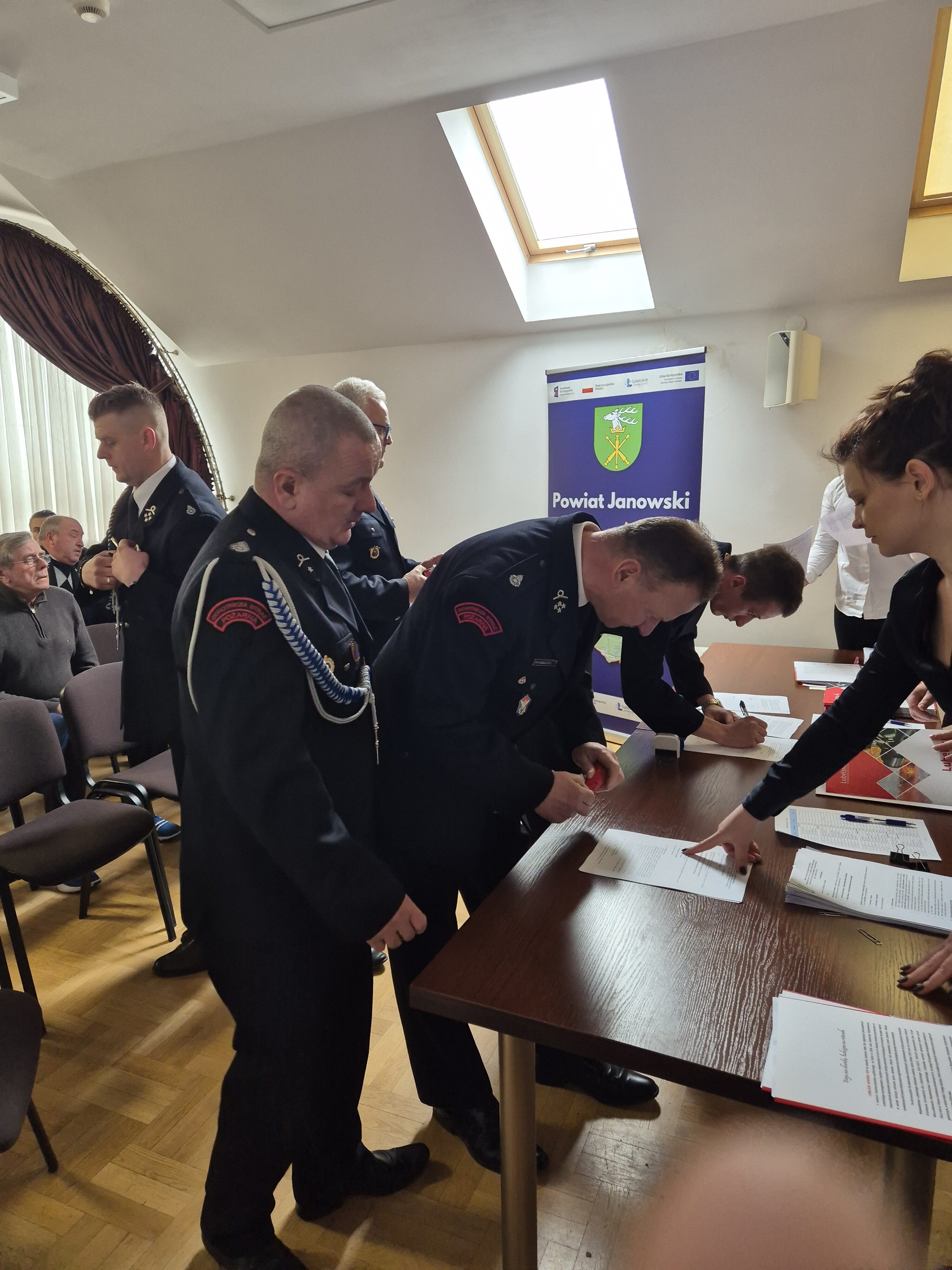 Grupa mężczyzn w mundurach strażackich podpisuje dokumenty przy drewnianym stole, w tle niebieski baner z herbem i napisem "Powiat Janowski".