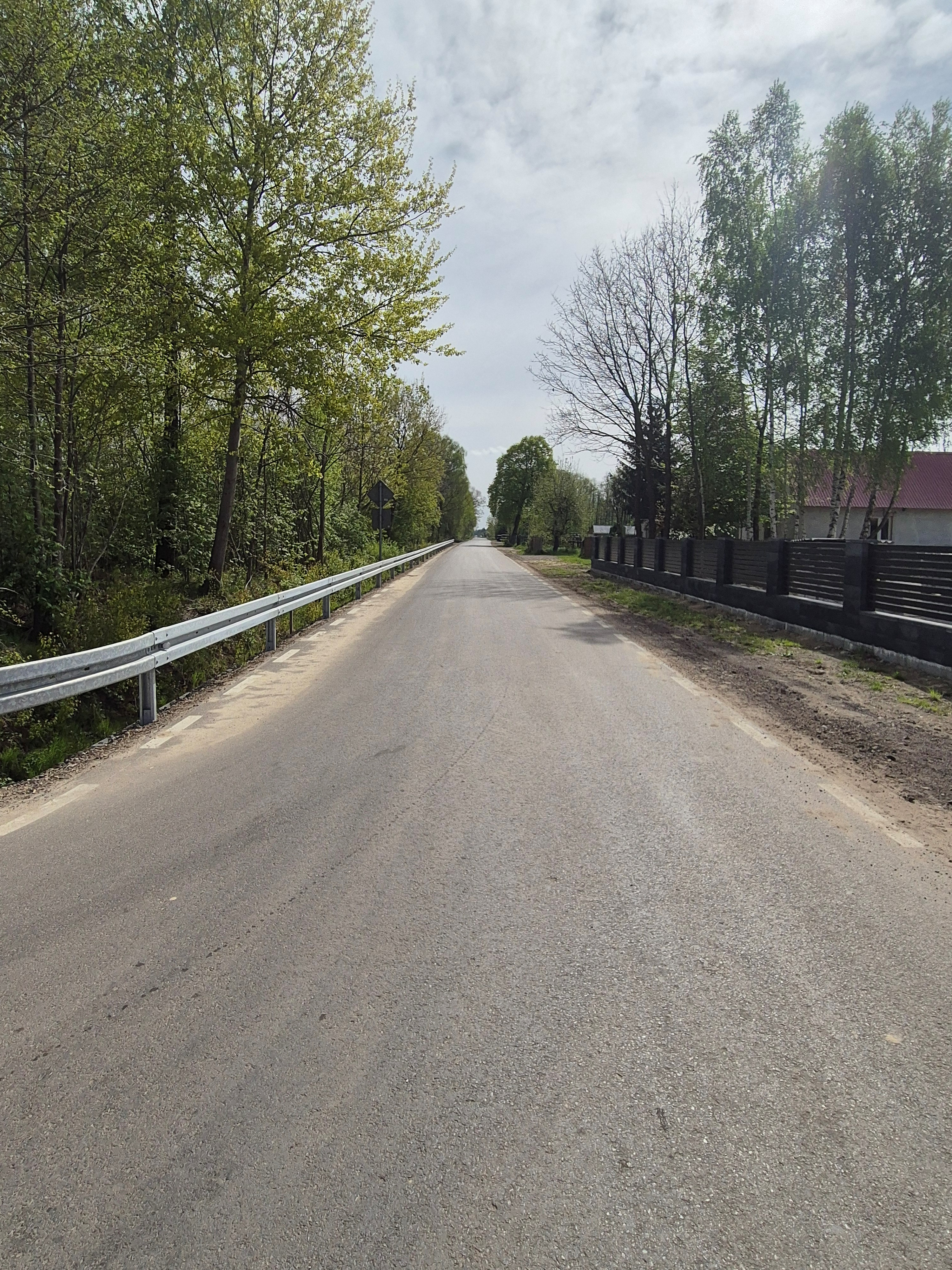 Prosta asfaltowa droga z metalową barierą po lewej stronie, po prawej czarne ogrodzenie i drzewa, niebo częściowo zachmurzone.