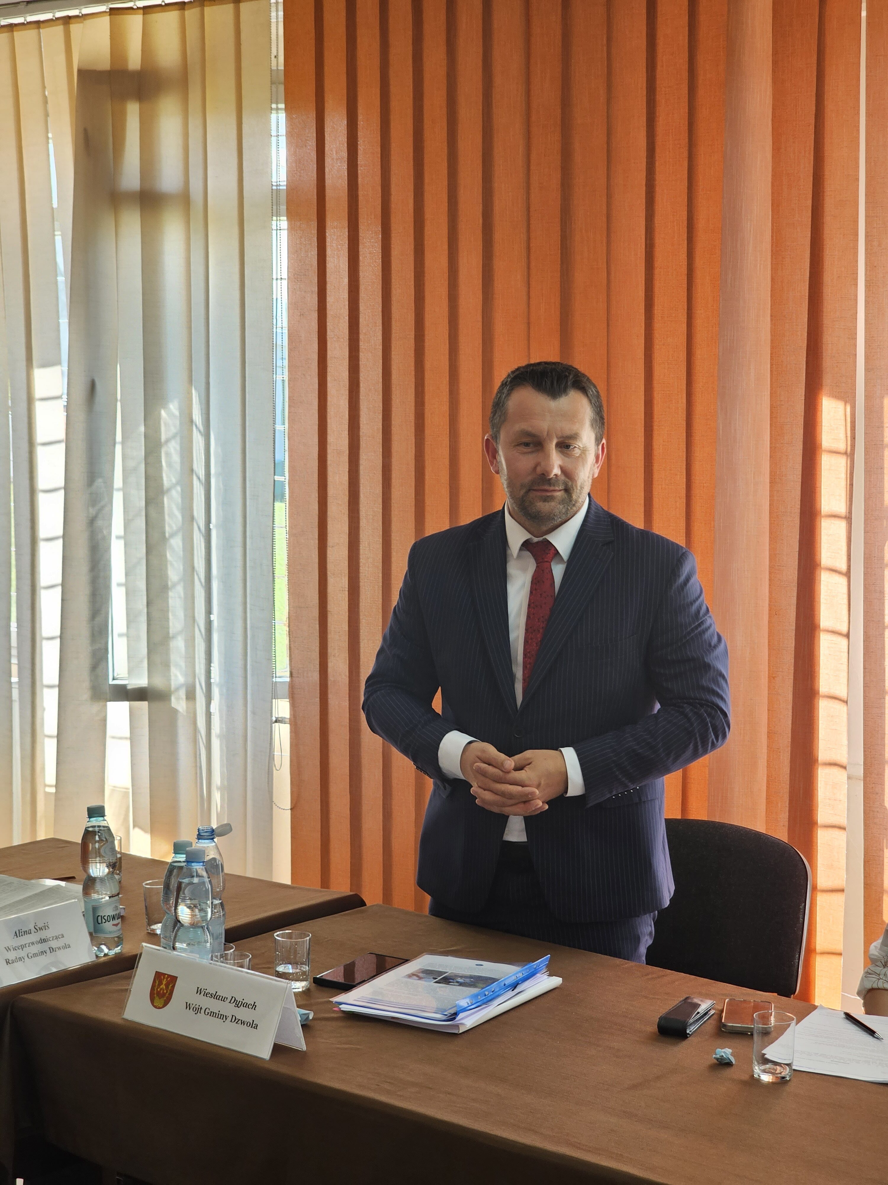 Mężczyzna w granatowym garniturze i czerwonym krawacie stoi przy stole z brązowym obrusem, na stole dokumenty, butelki wody i kartki, w tle pomarańczowo-beżowe zasłony.