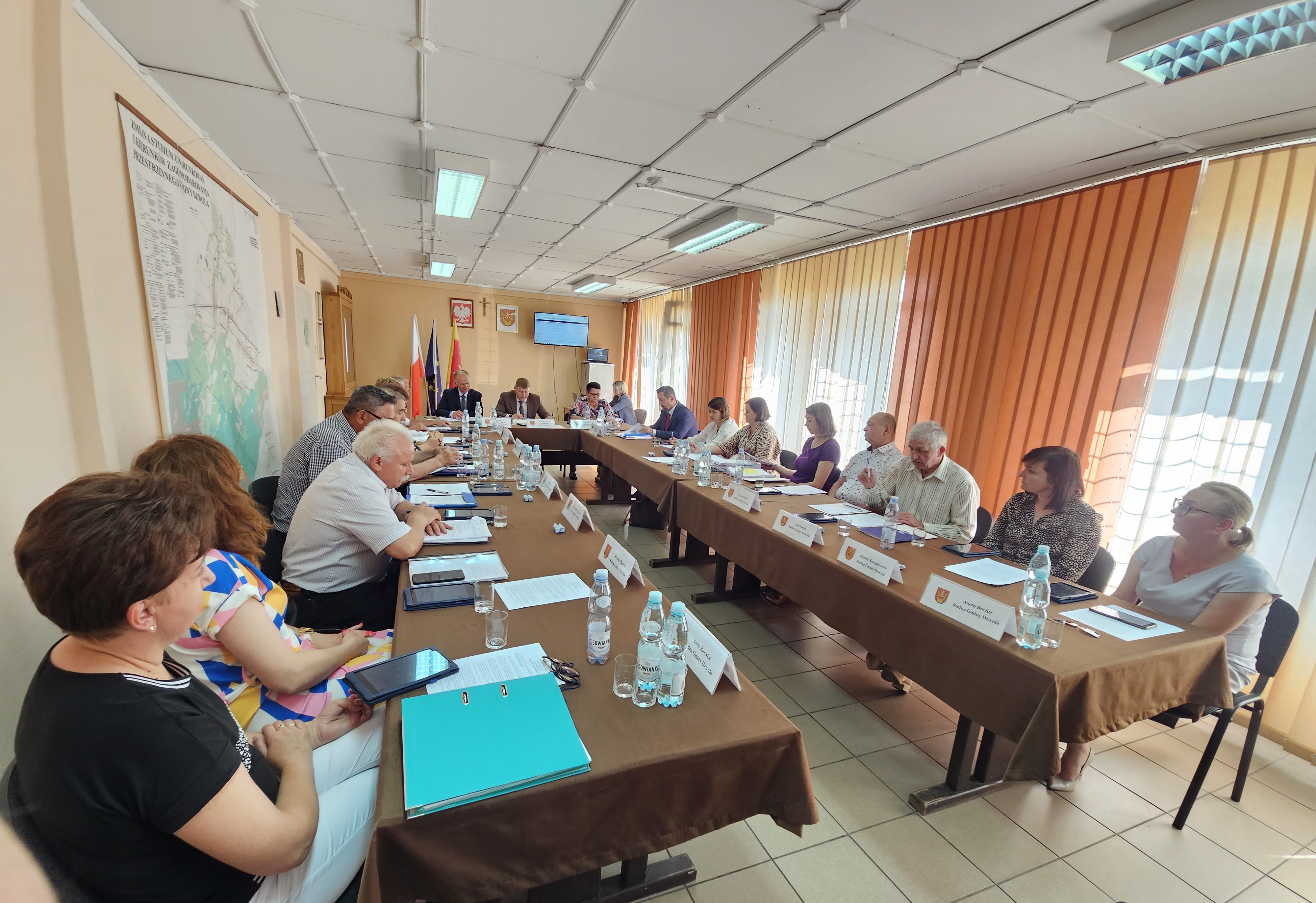 Ludzie siedzący przy długich stołach z brązowymi obrusami, plastikowe butelki i dokumenty, jasne zasłony, mapa na ścianie, flagi Polski i Unii Europejskiej, sufit z paneli.