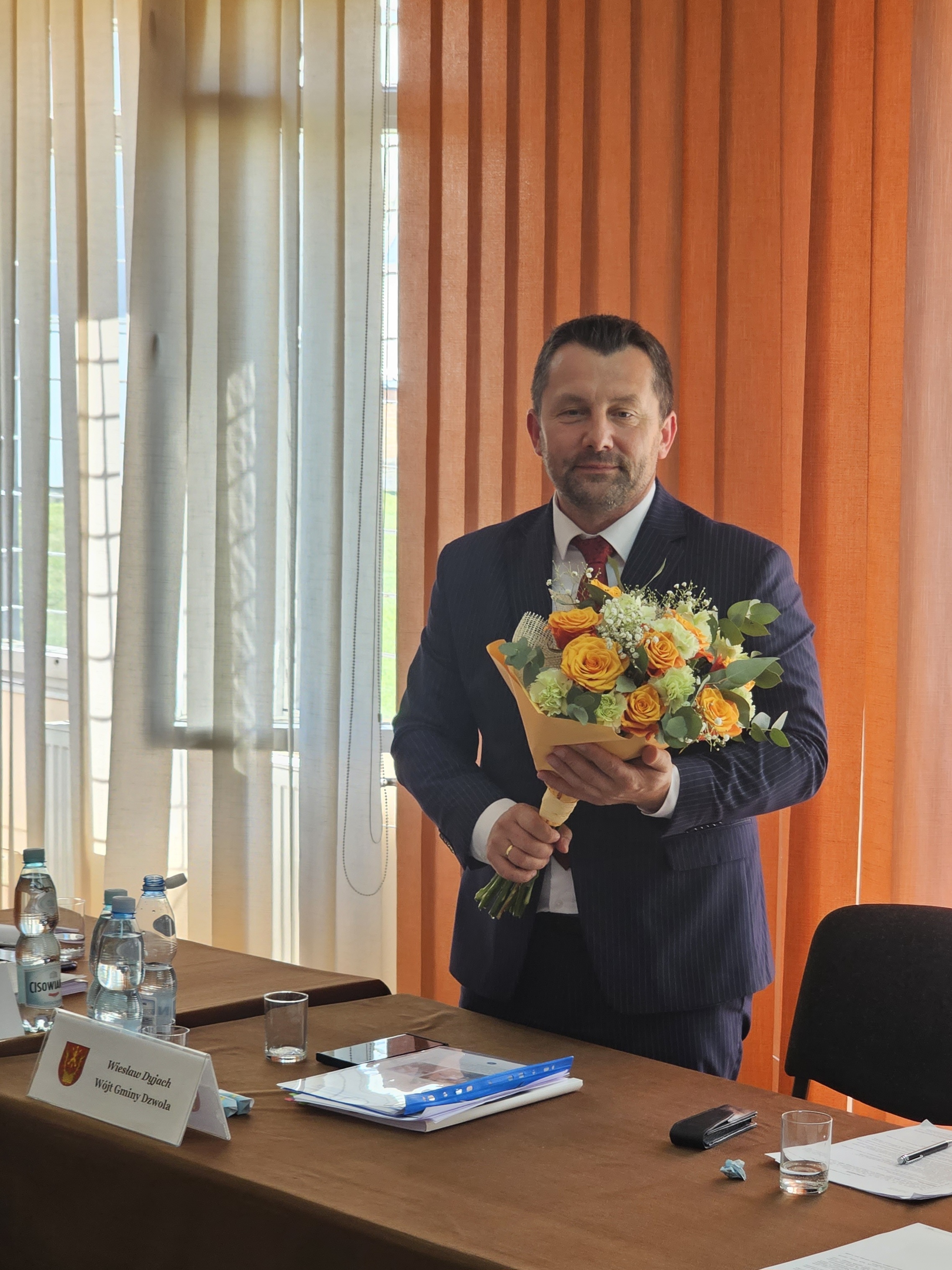 Mężczyzna w granatowym garniturze i czerwonym krawacie trzyma bukiet żółto-pomarańczowych kwiatów, stoi przy stole z dokumentami, butelkami wody i szklankami, w tle pomarańczowe i białe zasłony.