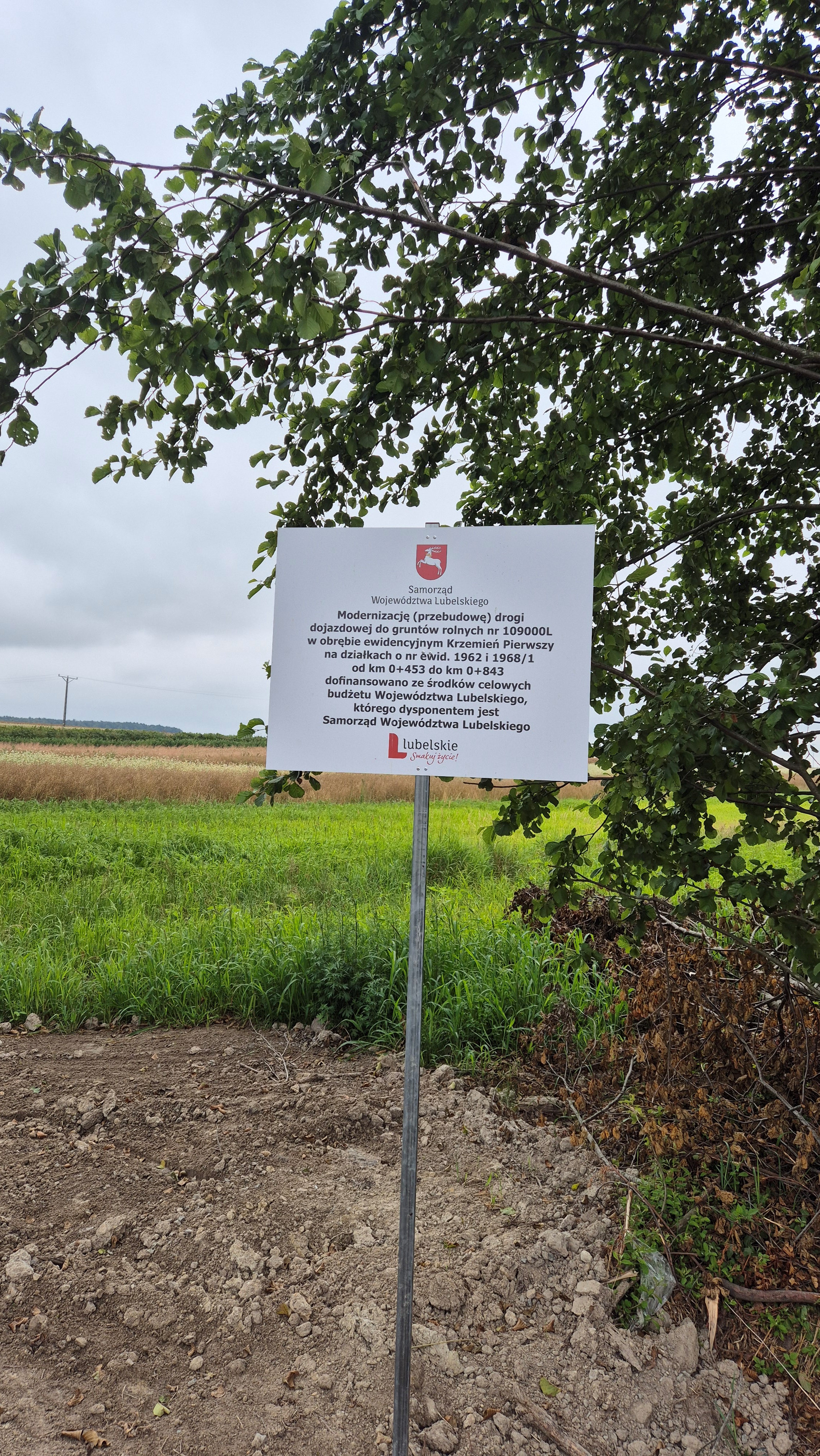 Tablica informacyjna na metalowej podporze na tle zielonej łąki, drzew z liśćmi i częściowo brązowej ziemi, niebieskie niebo z chmurami.