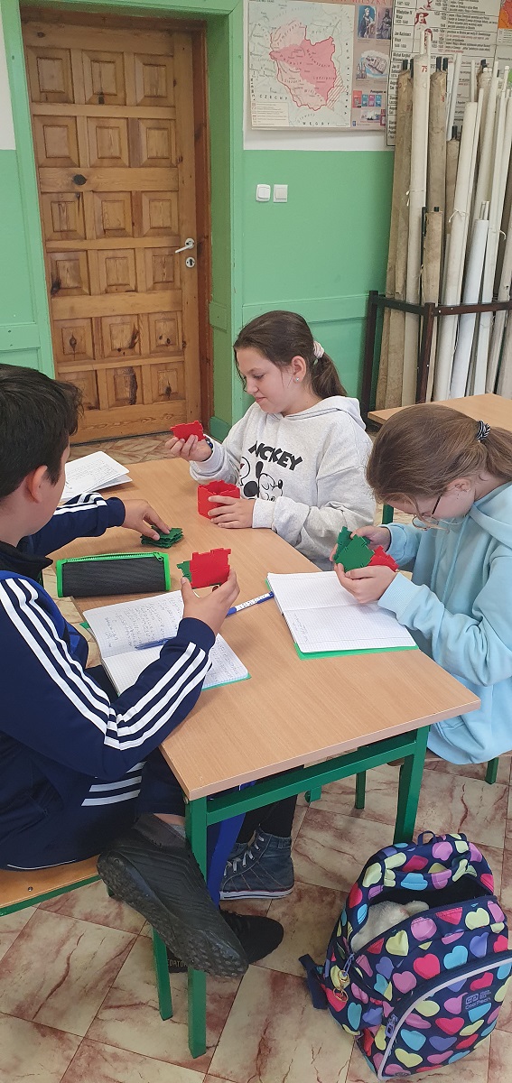 Trzy dzieci siedzą przy drewnianym stole w klasie, trzymają czerwone i zielone elementy puzzli, obok leżą zeszyty i piórnik, w tle drewniane drzwi i mapa na ścianie.