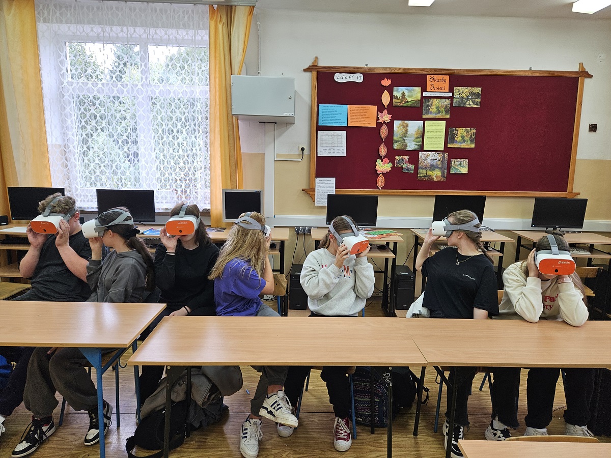 Siedem osób siedzi w klasie przy ławkach, każda ma na głowie biało-pomarańczowy zestaw VR, za nimi komputery, na ścianie bordowa tablica z kolorowymi kartkami i obrazami.