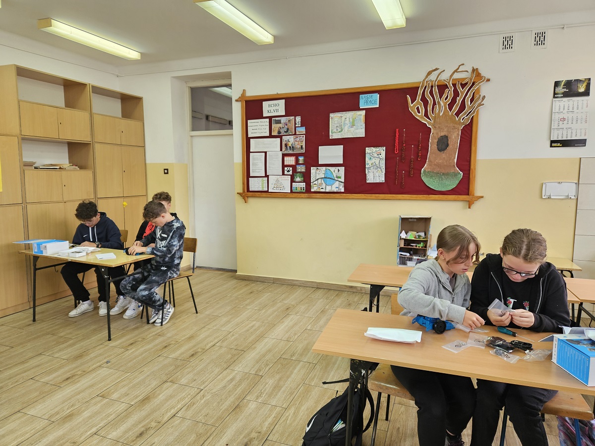 Wnętrze klasy szkolnej z czterema uczniami siedzącymi przy dwóch stołach, na ścianie tablica z czerwonym tłem i różnymi pracami plastycznymi, jasne meble i podłoga.