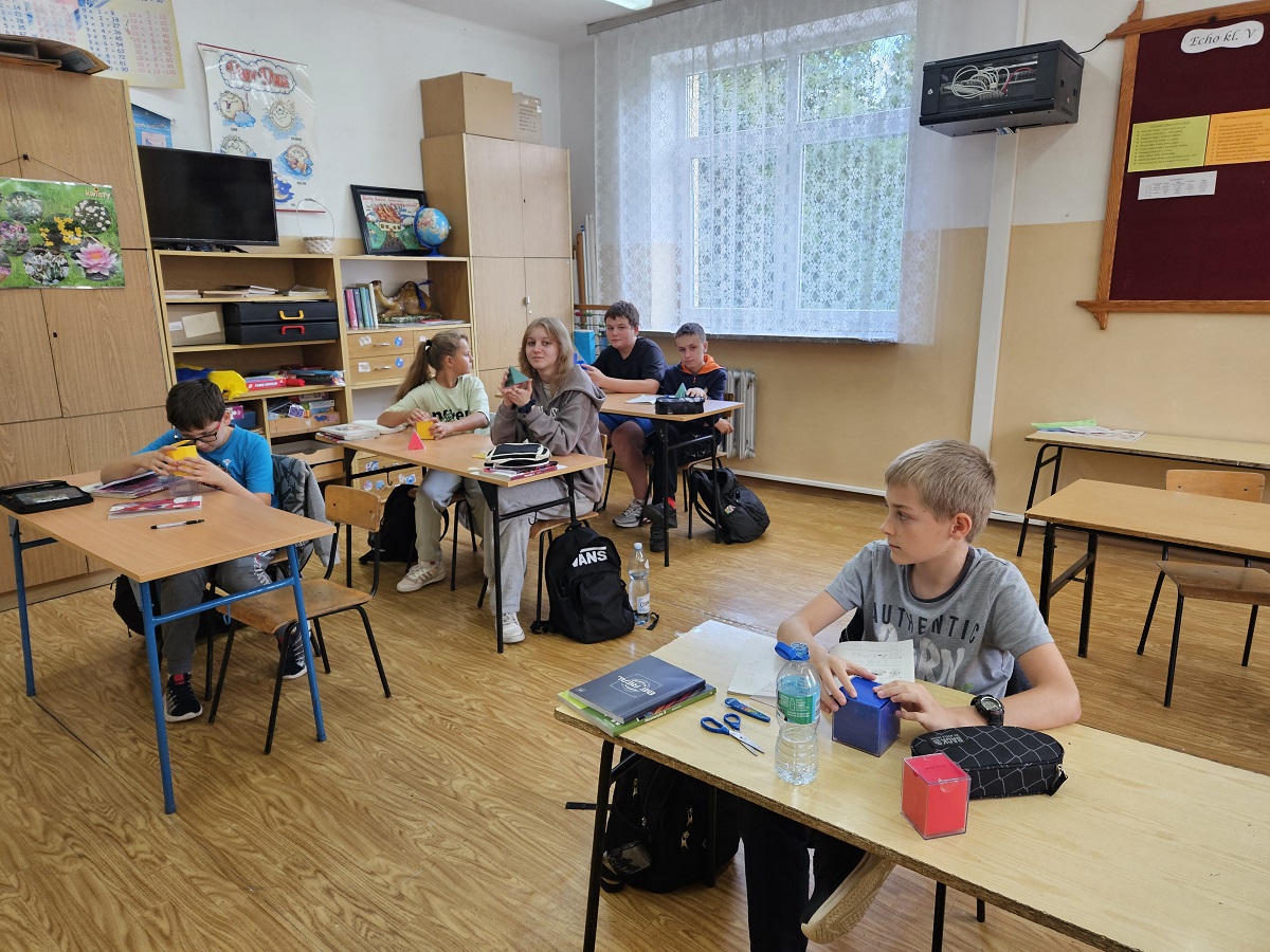 Klasa szkolna z sześcioma uczniami siedzącymi przy biurkach, drewniana podłoga, meble i tablica korkowa na ścianie, duże okno z białą firanką.