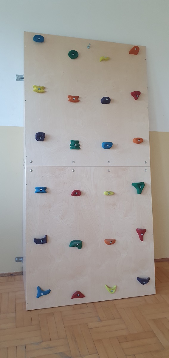 Drewniana ściana wspinaczkowa z kolorowymi chwytami w różnych kształtach, zamocowana na jasnej ścianie nad podłogą z drewnianych paneli.