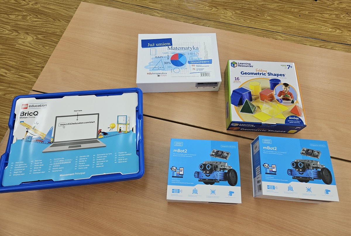 Zestawy edukacyjne na drewnianym stole, w tym LEGO BricQ Motion Prime, matematyka, kształty geometryczne i mBot2 w niebieskich i białych opakowaniach.