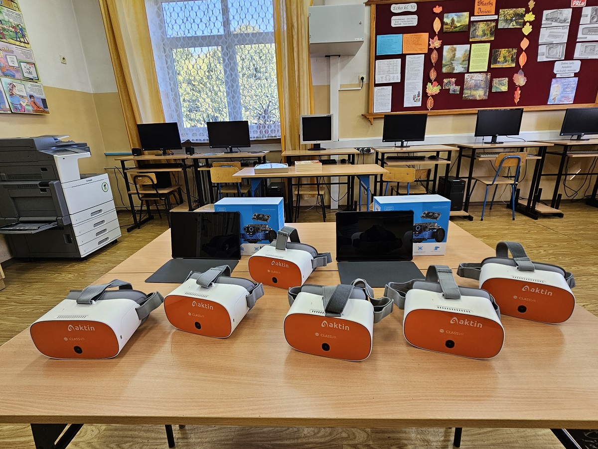 Sześć białych i pomarańczowych gogli VR na drewnianym stole, dwa tablety z czarnymi ekranami, niebieskie pudełka z obrazami robotów, w tle komputerowe stanowiska i tablica ogłoszeń.