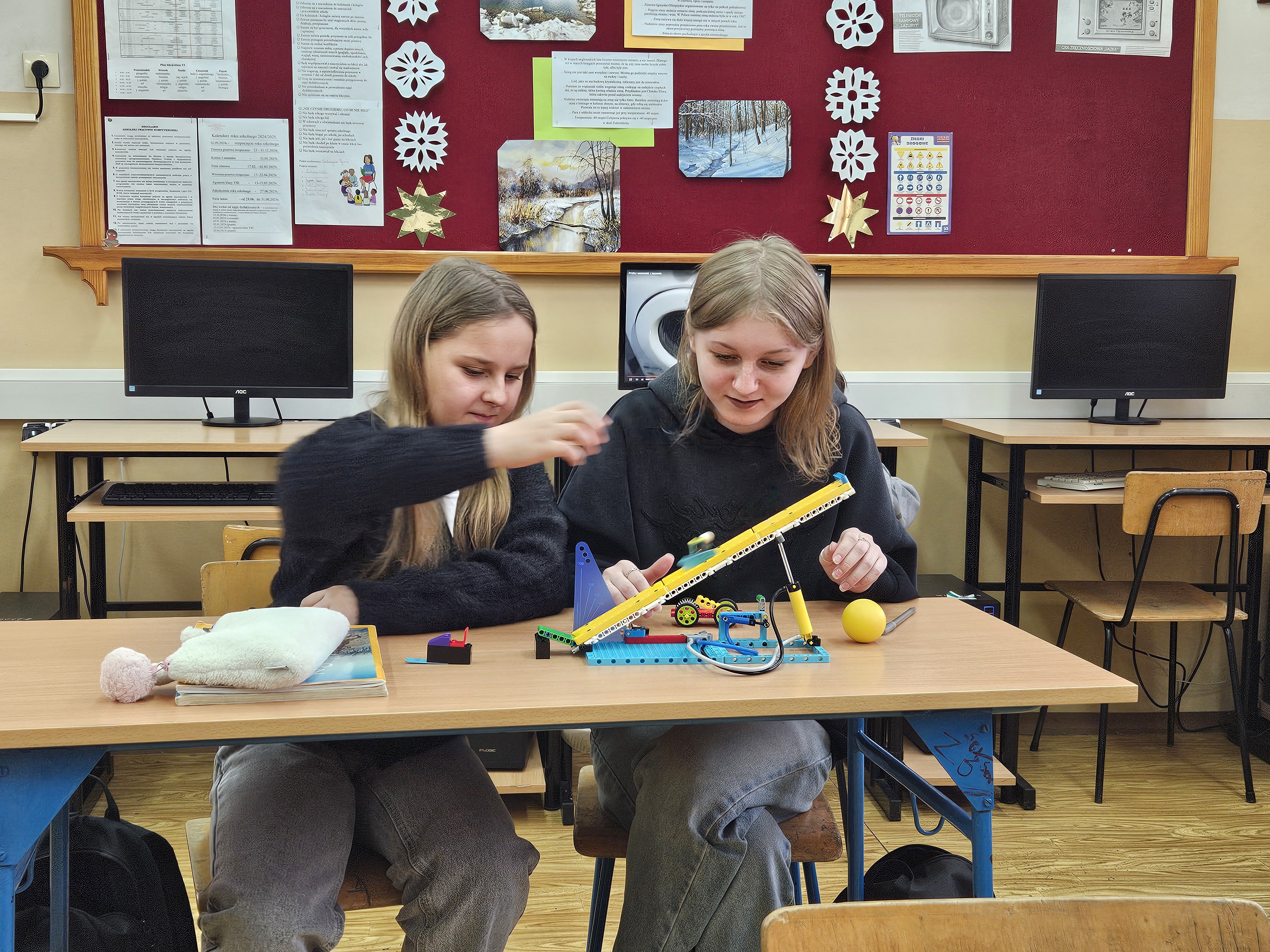 Dwie dziewczyny siedzą przy stole z modelem z klocków konstrukcyjnych, w tle tablica z kartkami i dekoracjami, za nimi trzy monitory komputerowe.
